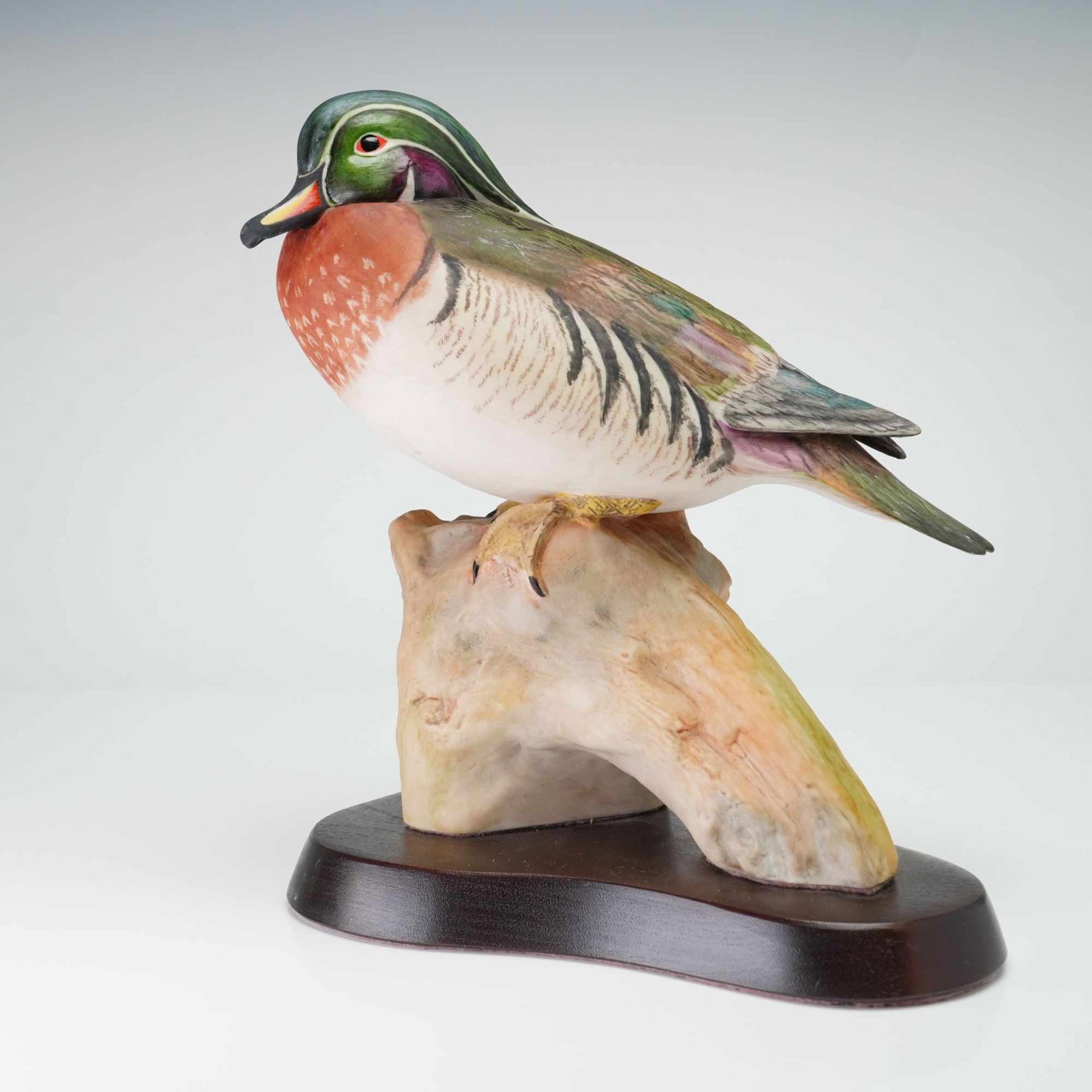 Cybis Porcelain Figurine on Wooden Base, Wood Duck 456: The bird is depicted in a naturalistic perching pose atop a piece of simulated driftwood. The figurine features the species characteristic iridescent green and purple crested head, white facial markin