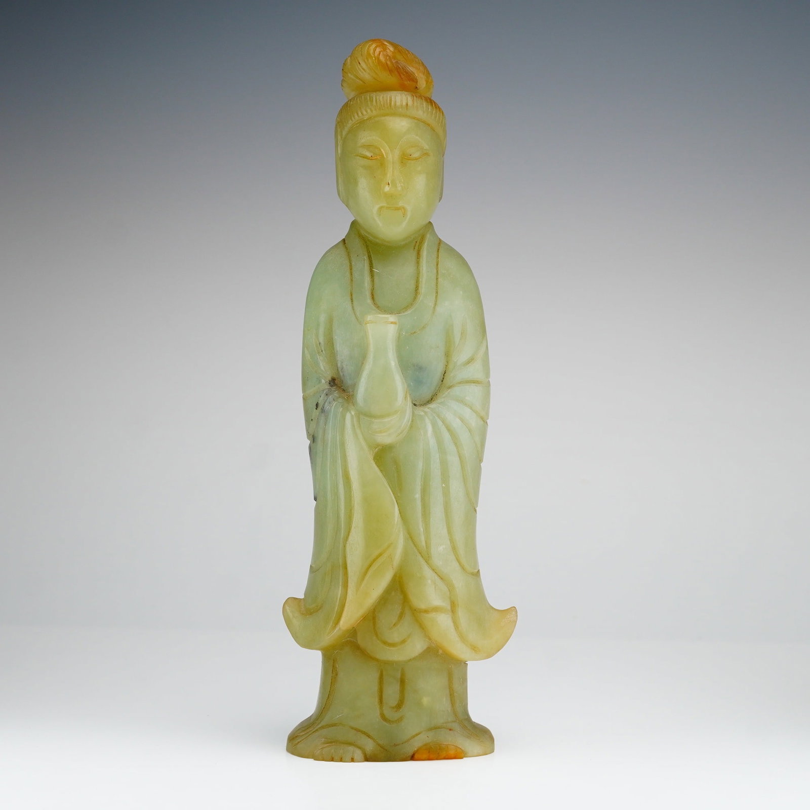 Chinese Carved Jade Figurine Depicting Guanyin: A Chinese carved nephrite figurine depicting Guanyin, the Goddess of Mercy. The deity is portrayed standing in traditional flowing robes with wide sleeves and flared hems, holding a small ritual
