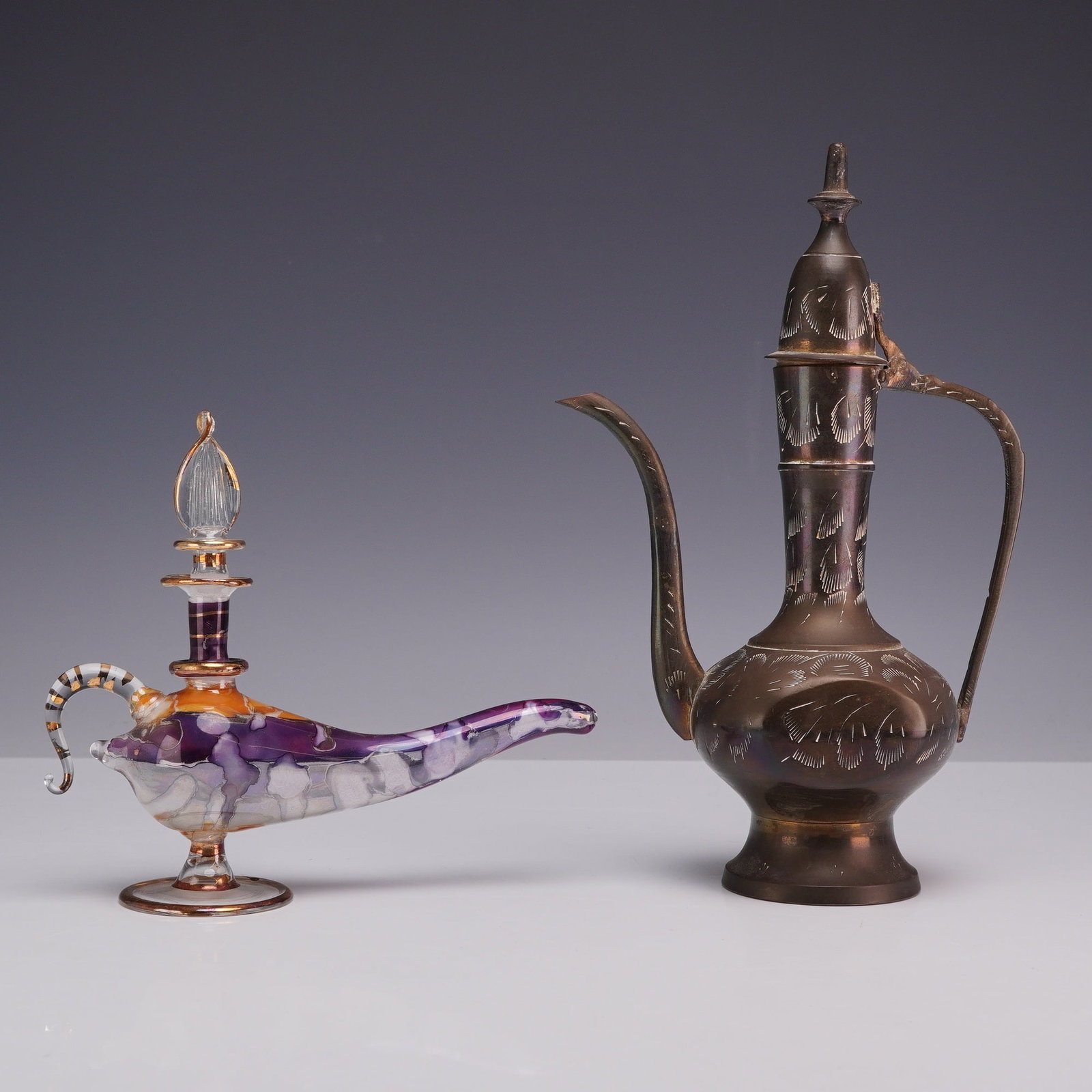 2pc Lidded Hinged Metal Ewer and Lamp Style Glass Perfume Bottle: This lot includes two decorative items: a lidded metal ewer and an Aladdin lamp-form glass perfume bottle. The ewer is constructed of brass with a dark patina and features hand-incised stylized leaf d