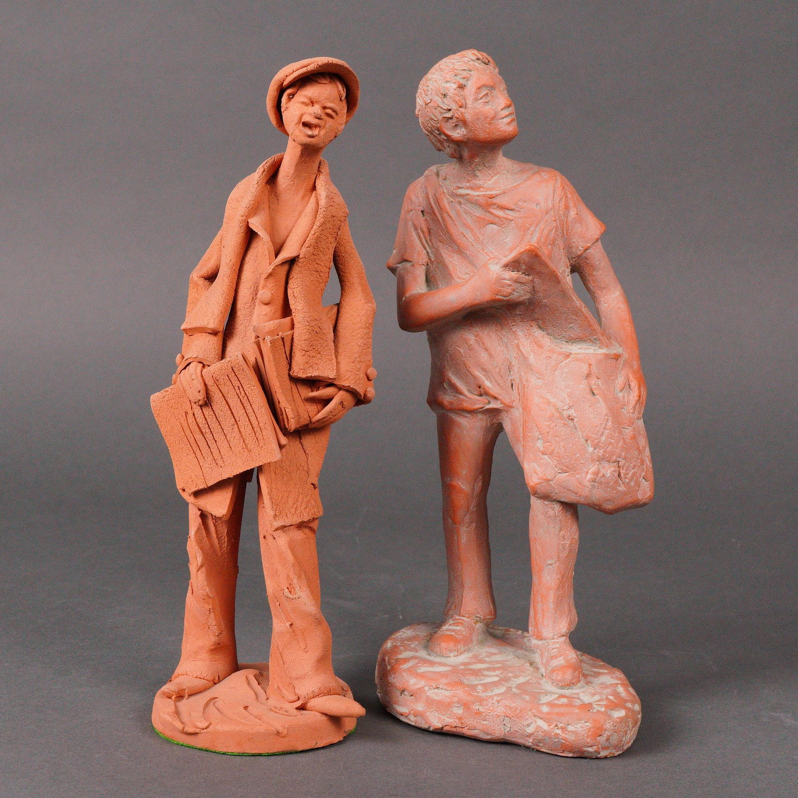 Group of Two Terracotta Newspaper Men Figurines: A group of two hand formed terracotta sculptures depicting elongated standing figures. One shows a person wearing a cap and long coat, holding newspapers and a book. The other presents a youth in a