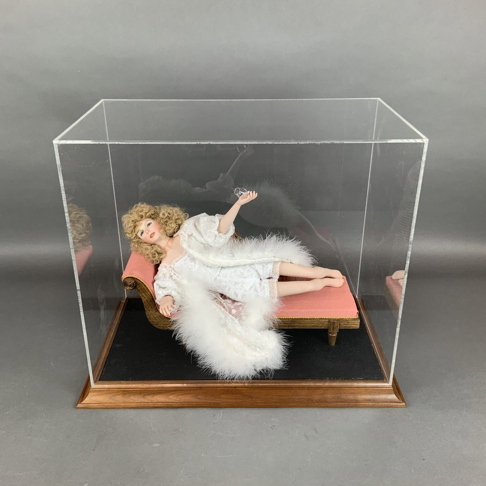 Bisque Porcelain Boudoir Doll Lace with Display Case: A boudoir-style collector doll depicted in a reclining pose on a miniature pink upholstered chaise longue. The head, hands, and feet are crafted from bisque porcelain, while the body is fabric-constru