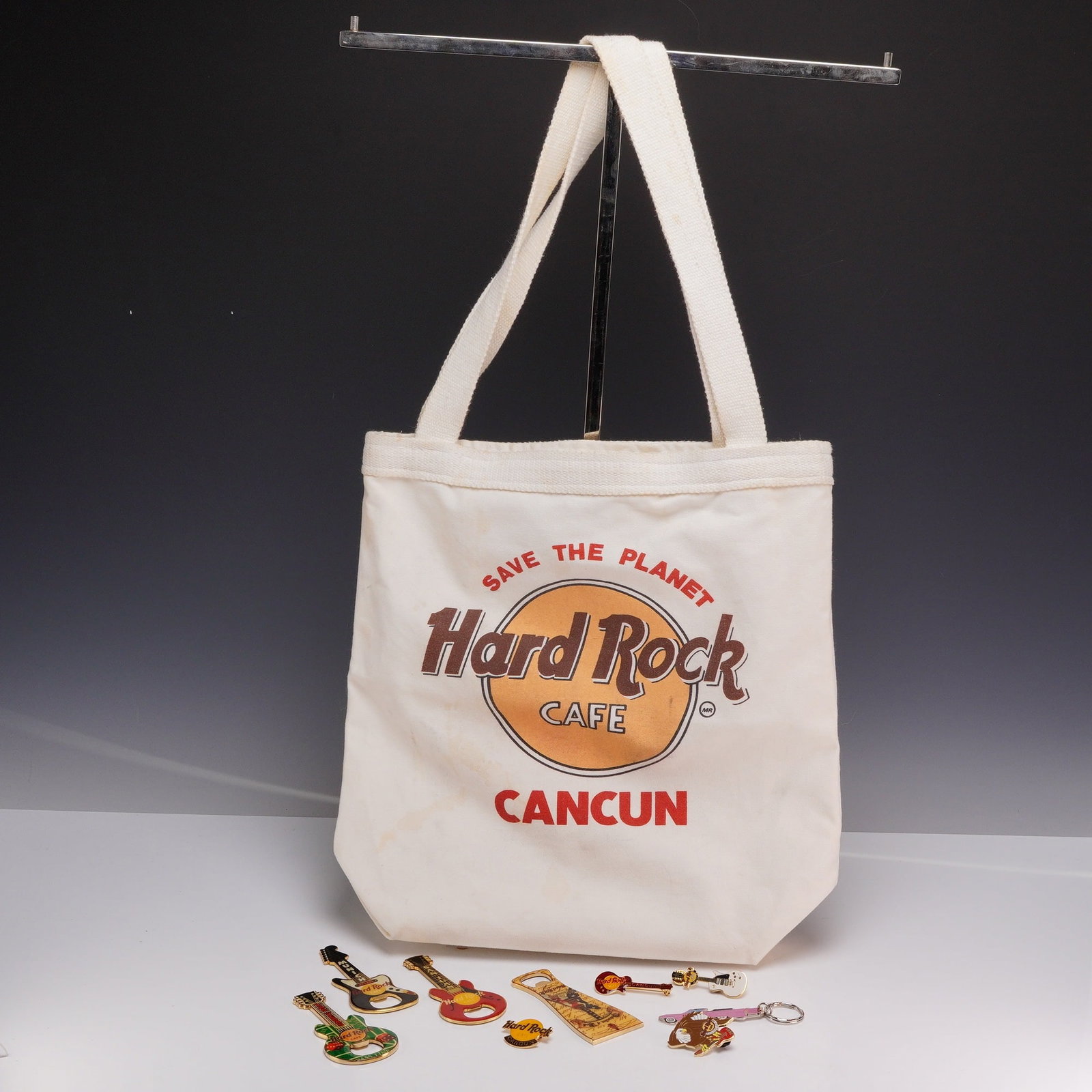 10pc Hard Rock Cafe international items including 1 tote bag, bottle openers and pins: A collection of ten Hard Rock Cafe international souvenir items consisting of one canvas tote bag and nine assorted accessories. The white canvas tote bag features a top zipper, two shoulder straps,