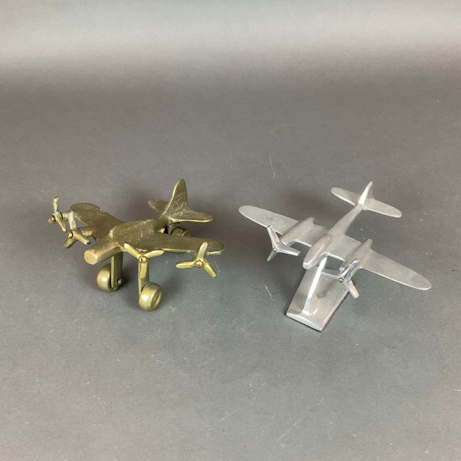 2pc Gold Tone and Silver Tone Metal Model Planes: This lot contains two metal plane decorative figures. The first model depicts a twin-engine propeller aircraft in a mid-century modern style, reminiscent of a Lockheed Electra. It is made of polished
