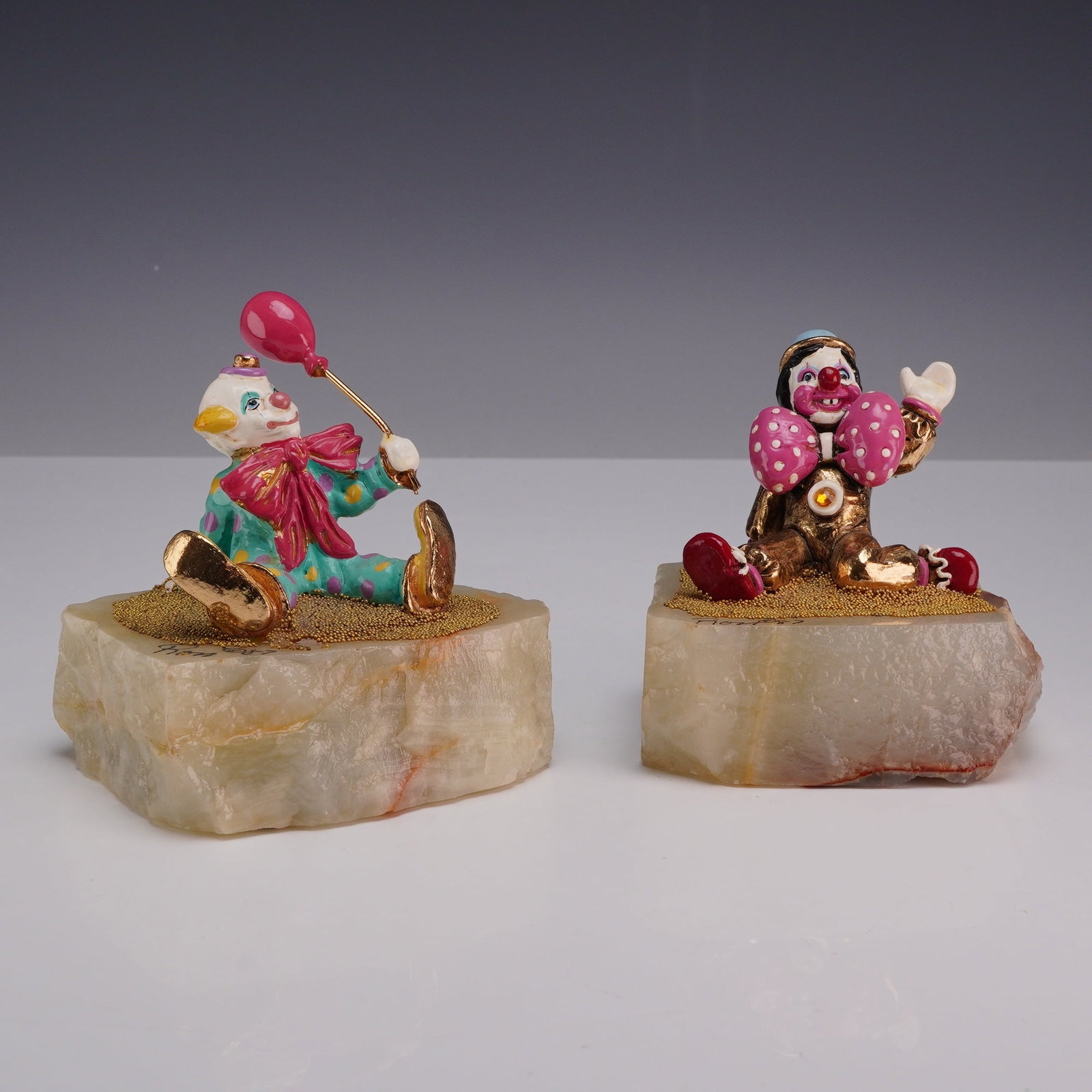 2pc Ronald A. Lee 24k Gold-Plated Clown Enamel Figurines: A lot of two hand-painted and 24k gold-plated clown figurines. The first figurine depicts a clown in a teal suit with multicolored polka dot decoration and a large pink bow, seated and holding a pink