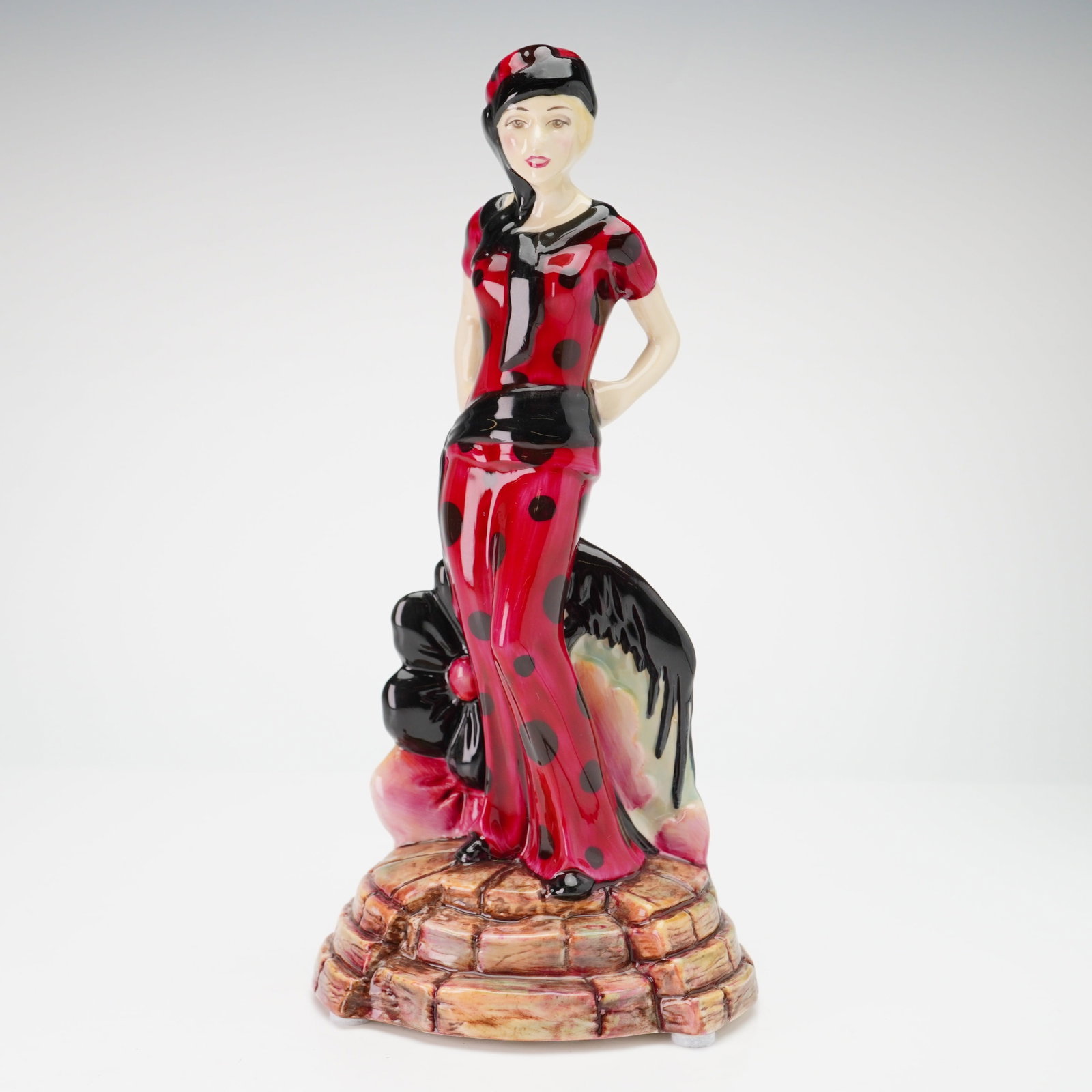 Kevin Francis Peggy Davies Ceramics Figurine Charlie Guild Piece 1998/1999: The design is executed in an Art Deco revival style, featuring a woman in a red outfit with large black polka dots and a matching headwrap, standing on a cobblestone-patterned base. The reverse of the