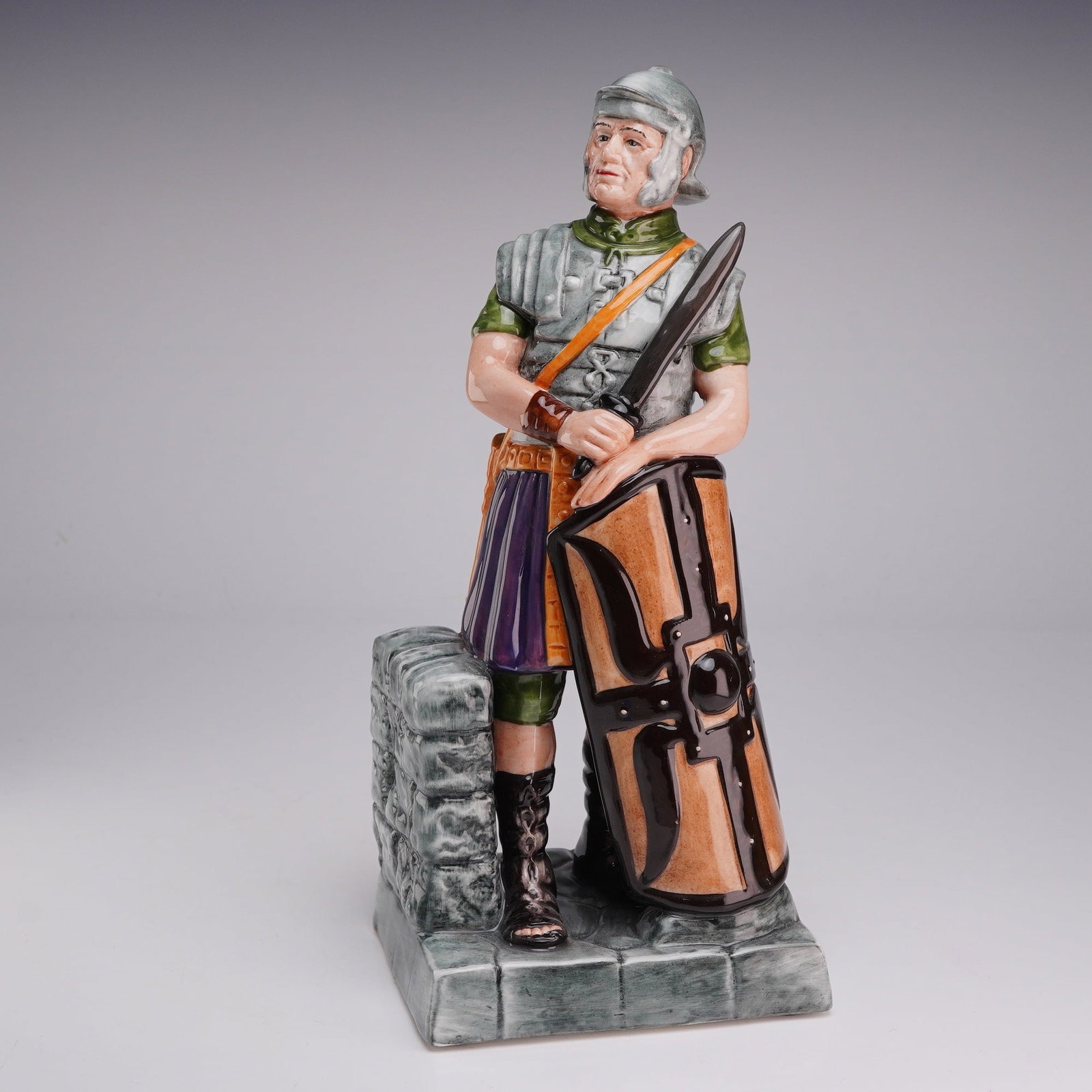 Royal Doulton Figurine The Centurion HN2726: This Royal Doulton glazed ceramic figurine depicts a Roman soldier in detailed military attire. The figure is depicted wearing a silver-toned helmet and lorica segmentata (banded armor) over a green t