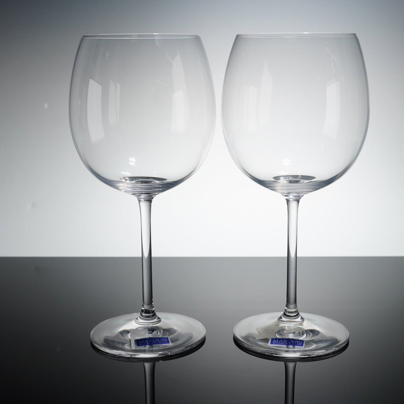 Pair of Marquis by Waterford Crystal Light Red Wine Glasses: Each glass features a large, rounded bowl atop a slender, elongated stem and a circular foot. The items are crafted from clear crystal and bear an etched MARQUIS BY Waterford hallmark on the underside
