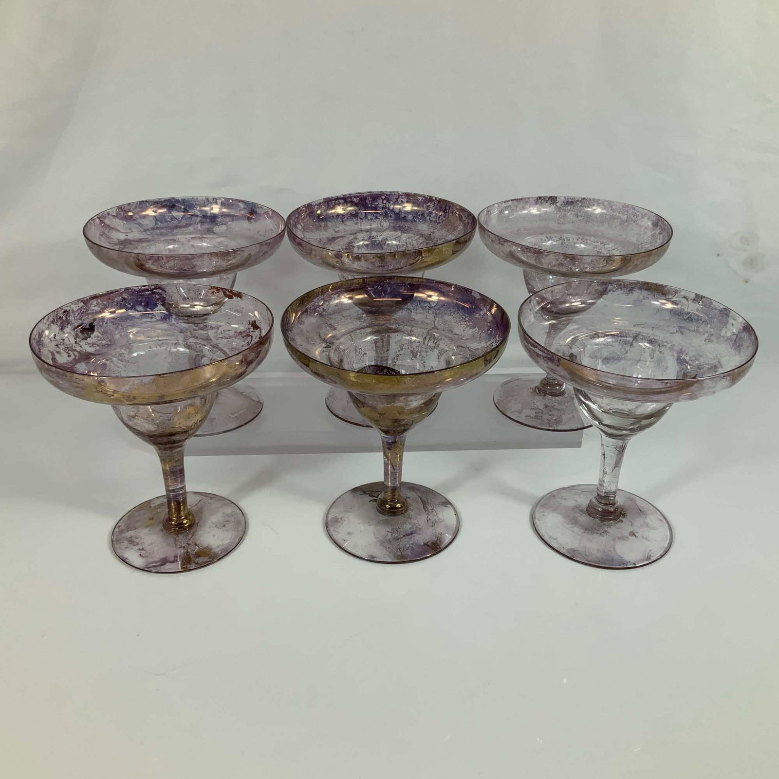 Set of Six Iridescent Marbled Purple Art Glass Margarita Glasses with Gold Toned Accents: This set of six margarita glasses is crafted from artisan art glass featuring wide, shallow bowls and stepped stems. The glasses are decorated with a mottled iridescent finish in shades of purple and