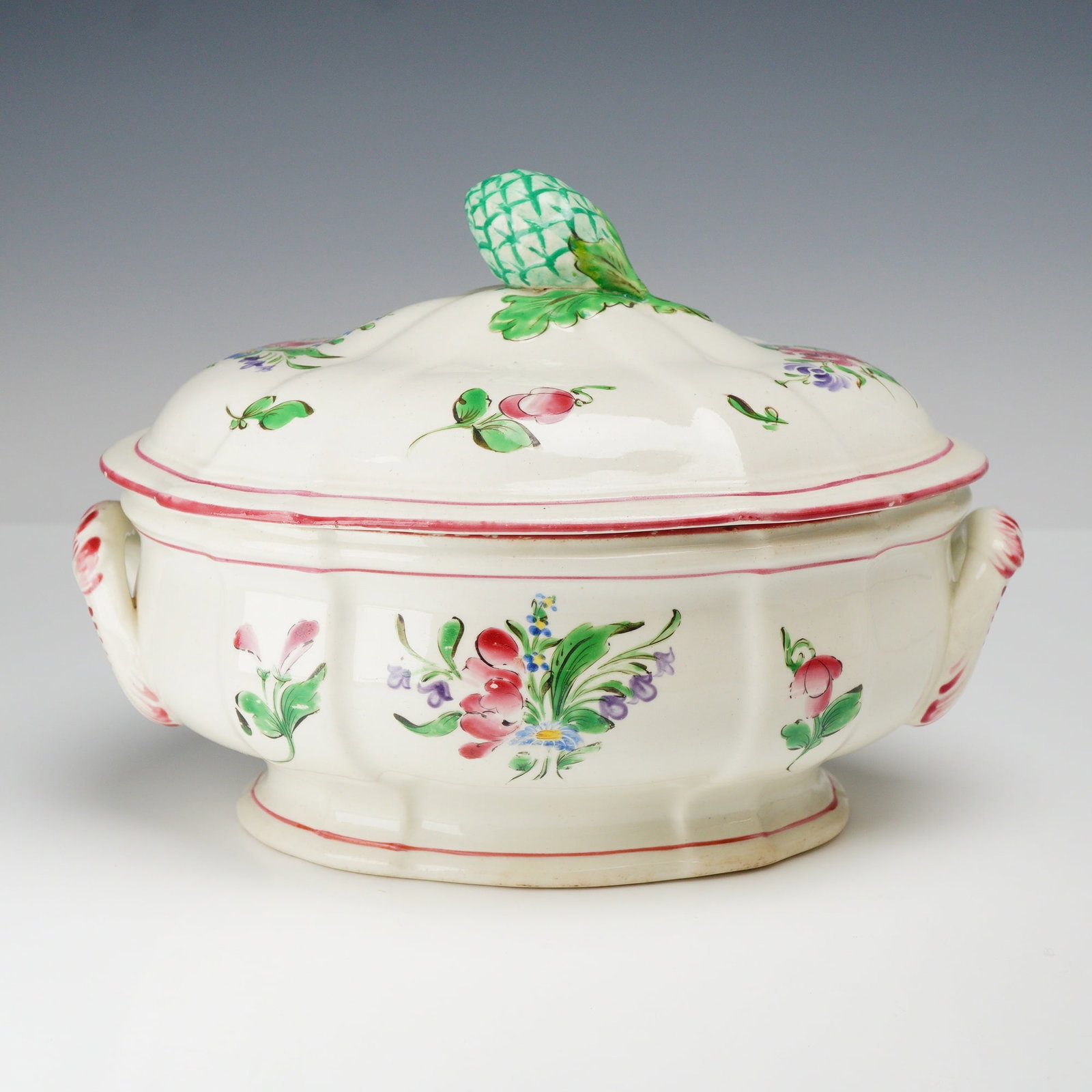 K and G Luneville Faience Lidded Soup Tureen with Artichoke Finial: A Keller and Guerin (K and G) Luneville hand-painted faience soup tureen and lid. The earthenware piece features an oval, lobed body supported by a scalloped pedestal foot. Two integrated scroll