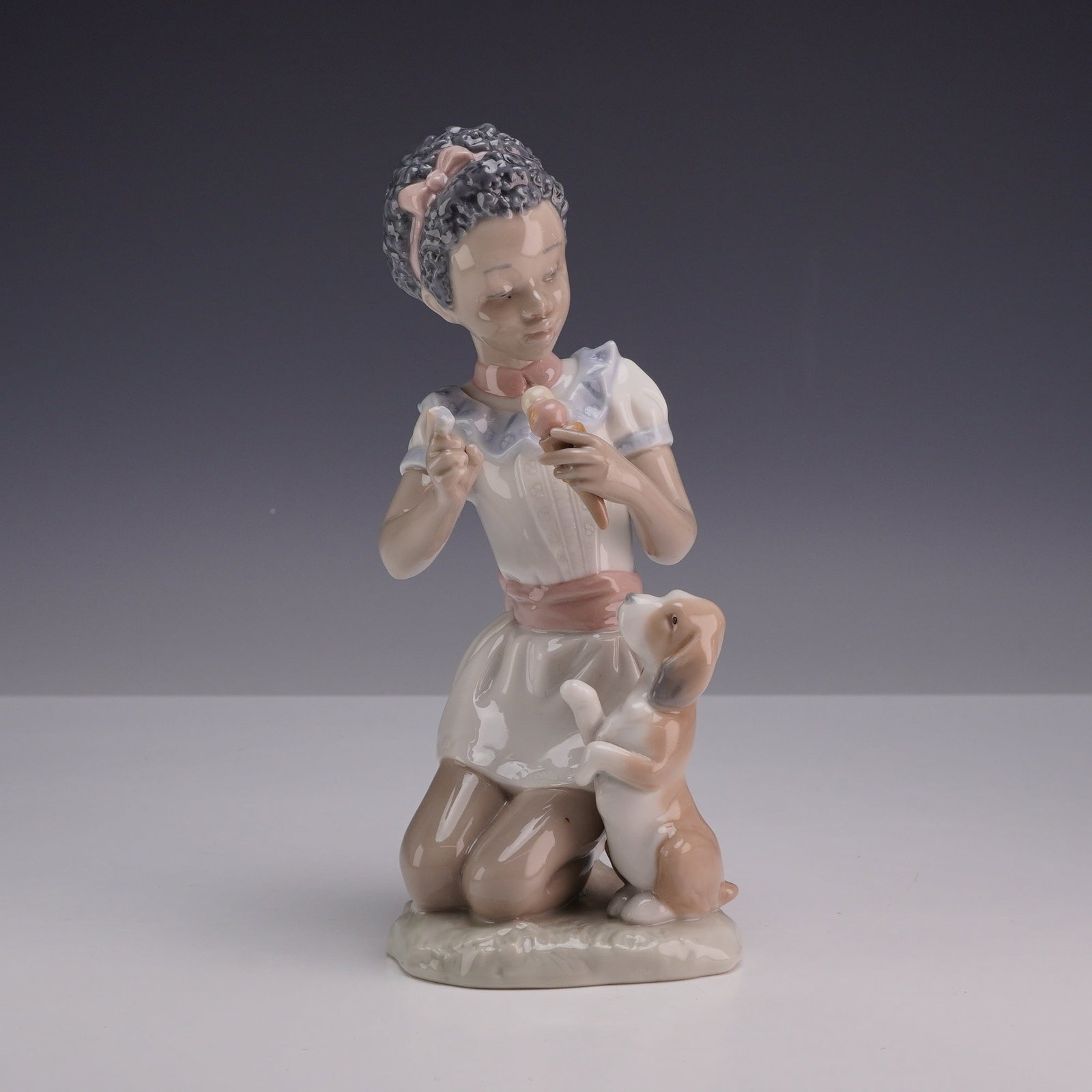 Lladro Porcelain Figurine Sharing Sweets 01005836: The figurine depicts a young girl with a dark skin tone kneeling while holding a pink ice cream cone. A small brown and white puppy is shown jumping up toward her. The girl wears a white dress with a 