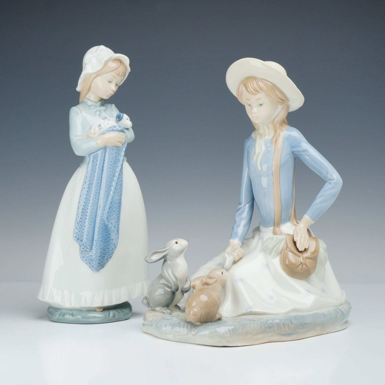 2pc Nao by Lladro Porcelain Girl Figurines: This lot features two Nao by Lladro porcelain figurines, including a girl feeding rabbits and a girl holding a puppy. The first figurine shows a standing girl in a blue bodice and bonnet cradling a sm