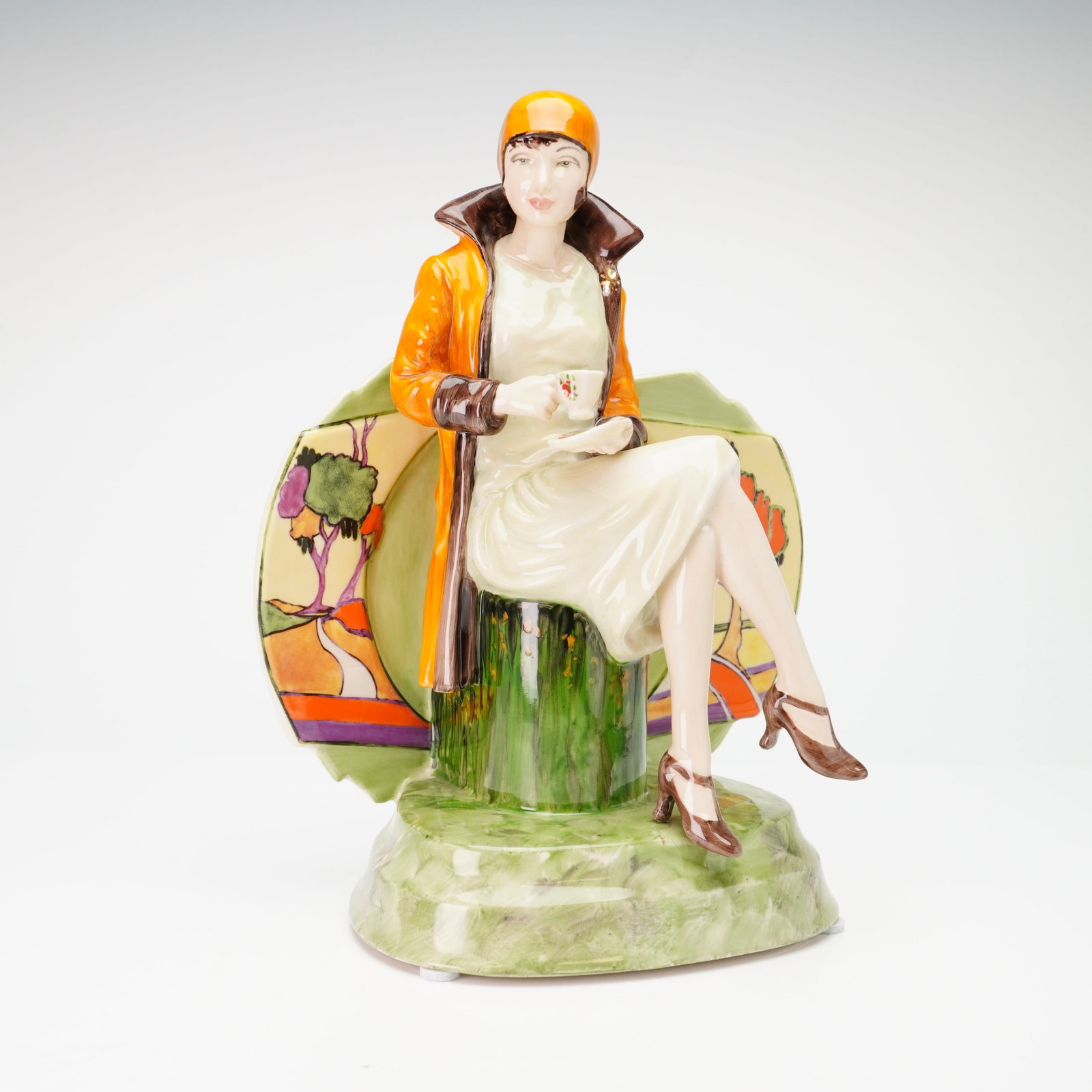 Peggy Davies Limited Edition Ceramic Figurine, Afternoon Tea: This hand-painted sculpture depicts a woman in 1920s attire seated on a green-glazed tree stump holding a teacup. The figure wears an orange cloche hat and a long orange coat with dark brown trim over