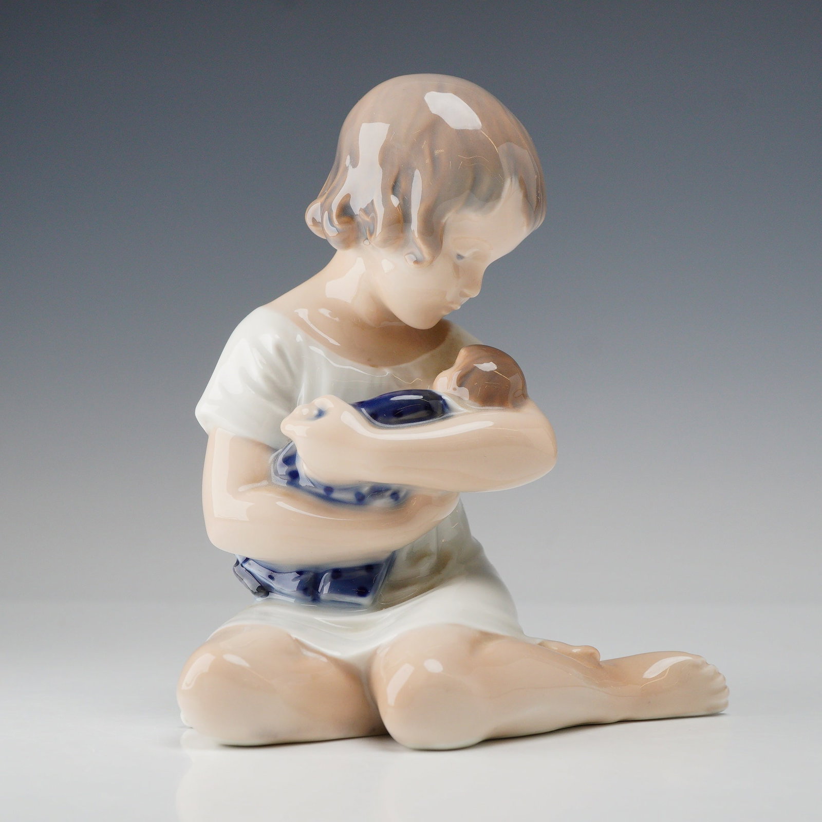 Royal Copenhagen Porcelain Figurine, Girl with Doll 1938: Royal Copenhagen porcelain figurine, model number 1938, portraying a young girl seated on the ground cradling a doll. The figure is barefoot and wears a short-sleeved white garment, gazing down at the