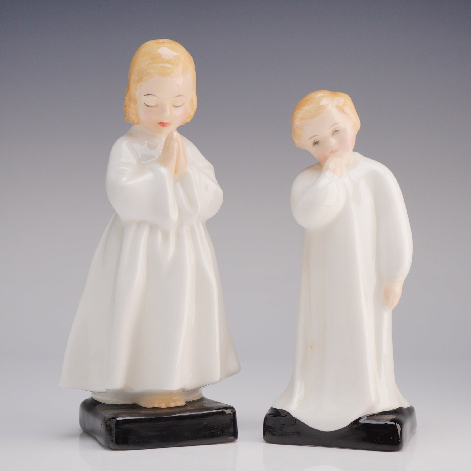 2pc Royal Doulton Figurines Bedtime HN 1978 and Darling HN 1985: The 2pc set depicts young children, modeled in white robes, standing on square black-glazed bases. The figure "Bedtime" portrays a girl with blonde hair and eyes closed, hands clasped in prayer. The f