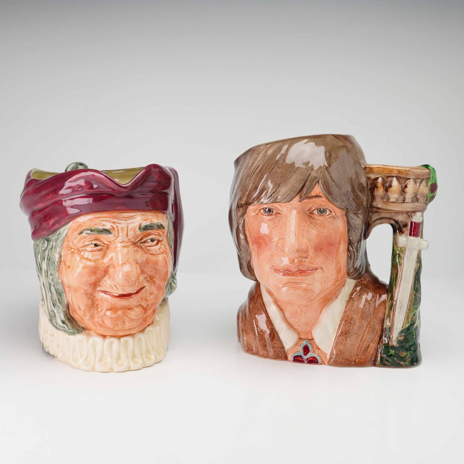 2pc Royal Doulton Character Jugs Simon the Cellarer D5504 and Romeo D6670: This lot consists of two glazed earthenware character jugs manufactured by Royal Doulton. The first jug is Simon the Cellarer, model D5504, depicting an elderly man with a ruddy complexion, wearing a 