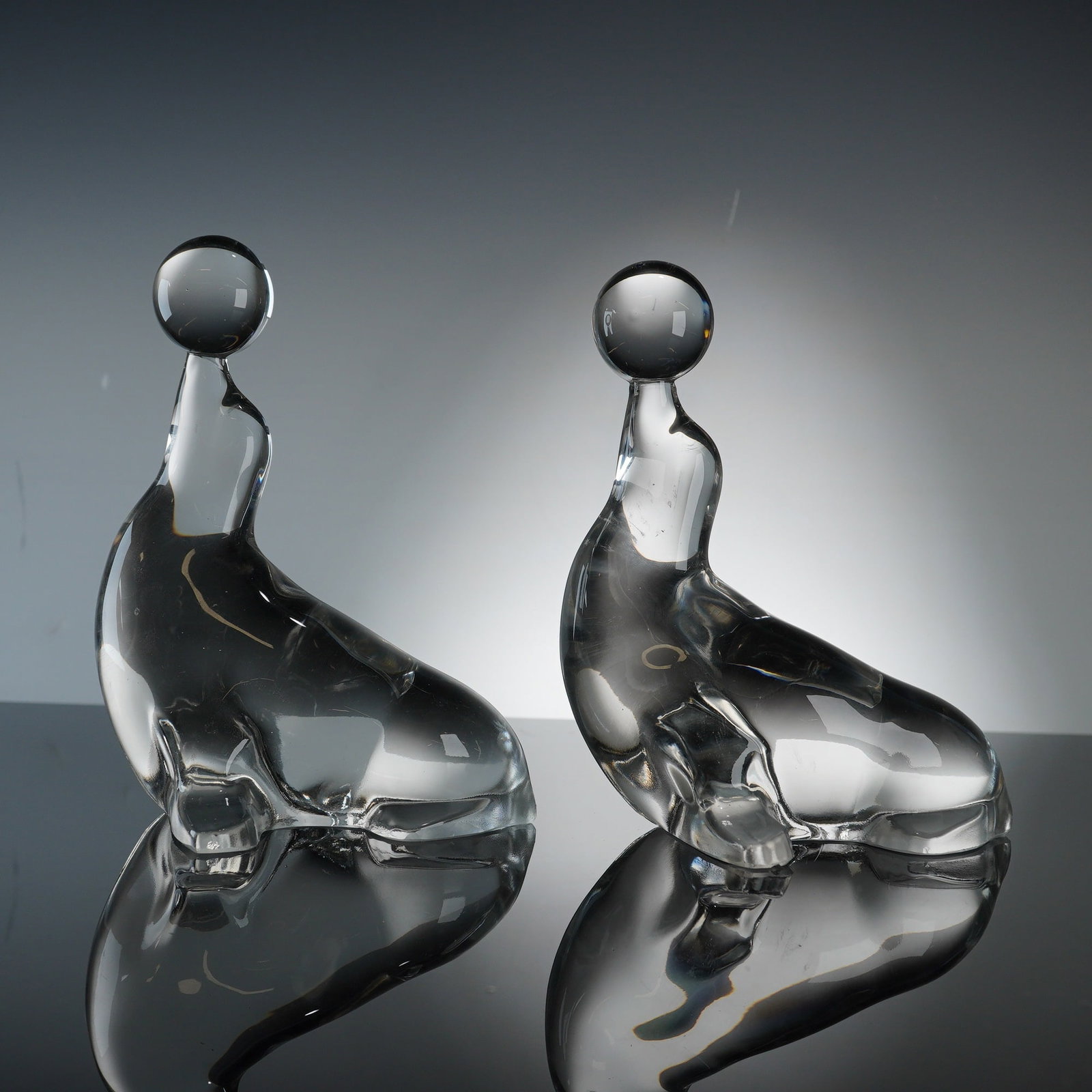 2pc Clear Glass Seal Figures w/ Balls Balanced on Noses: This set of two seal figurines is depicted with a ball balanced on its nose, modeled in a stylized, upright pose with an arched back and head tilted upward. The pieces are crafted from solid,