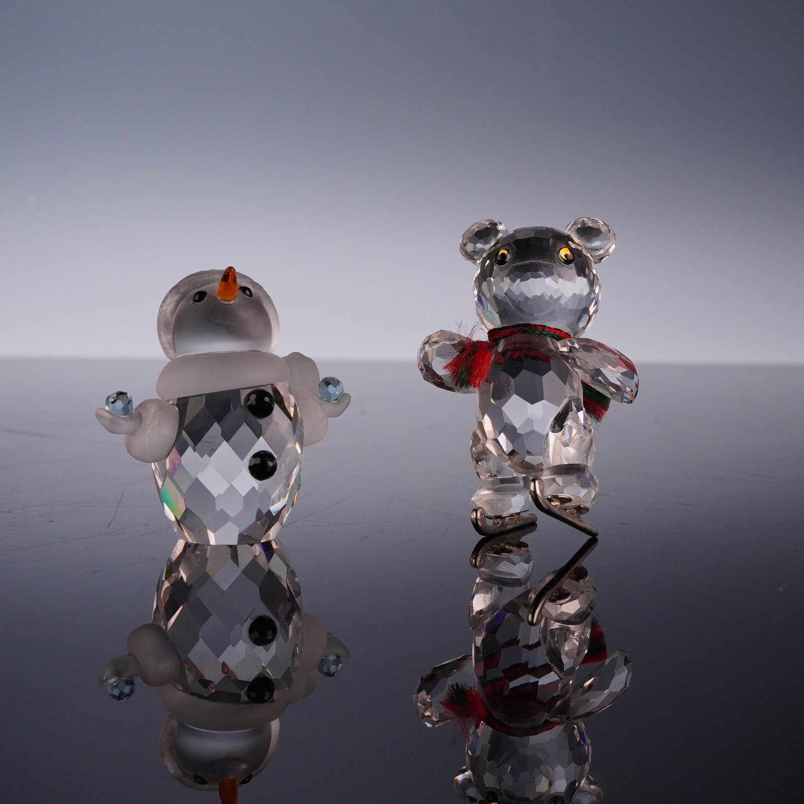 2pc Swarovski Crystal Figurines Including Kris Bear Ice Skates and Little Snowman: A set of two Swarovski cut lead crystal figurines from the holiday collection. The first piece is the Kris Bear Ice Skating, featuring a faceted clear crystal body, yellow and black eyes, and a red