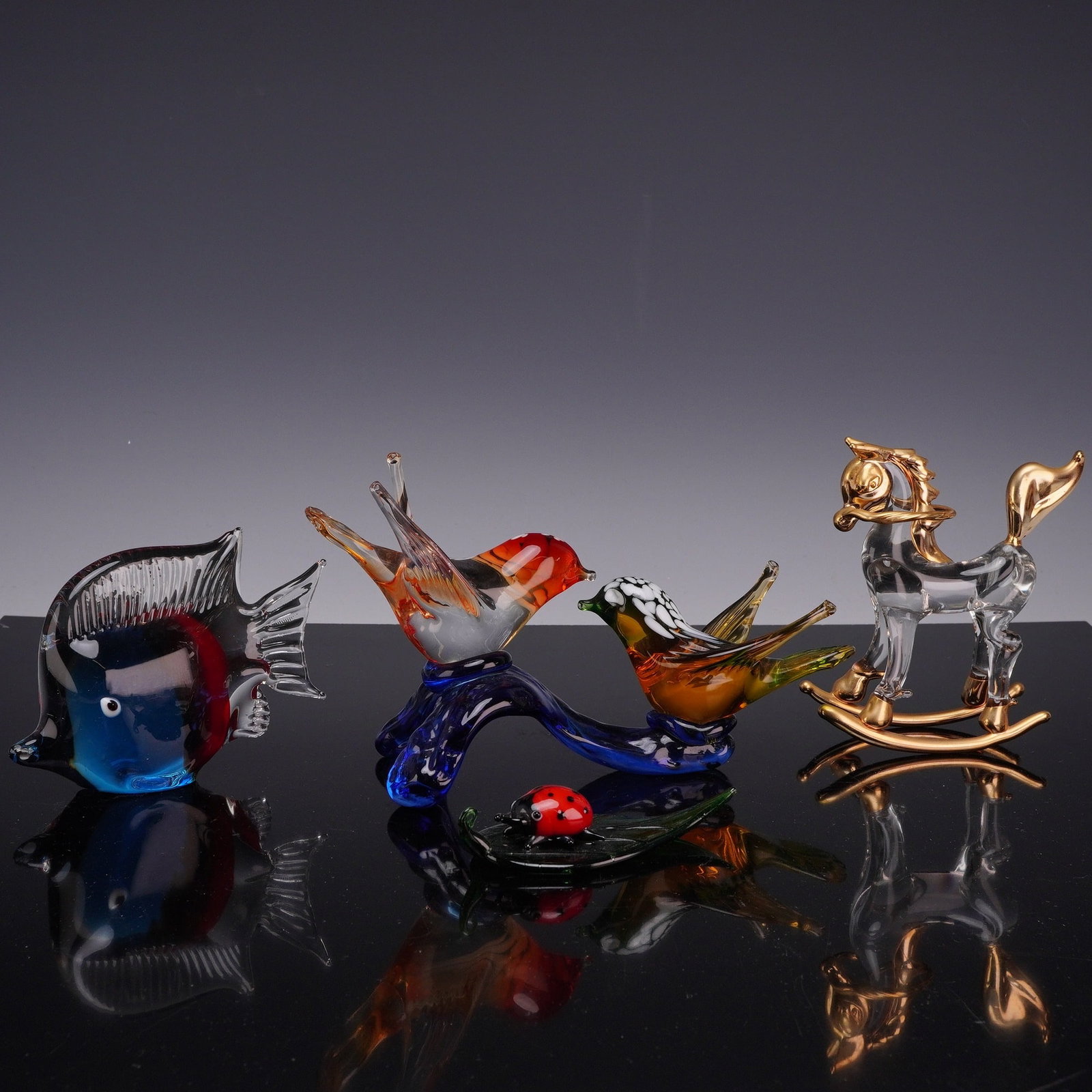 4pc Blown Glass Animals Including Fish, Rocking Horse, Ladybug on Leaf, and Two Birds on Branch: This collection consists of decorative glass figurines in various styles and techniques. Included is a polychrome hand-blown tropical fish with deep blue and red glass encased in clear glass, featurin