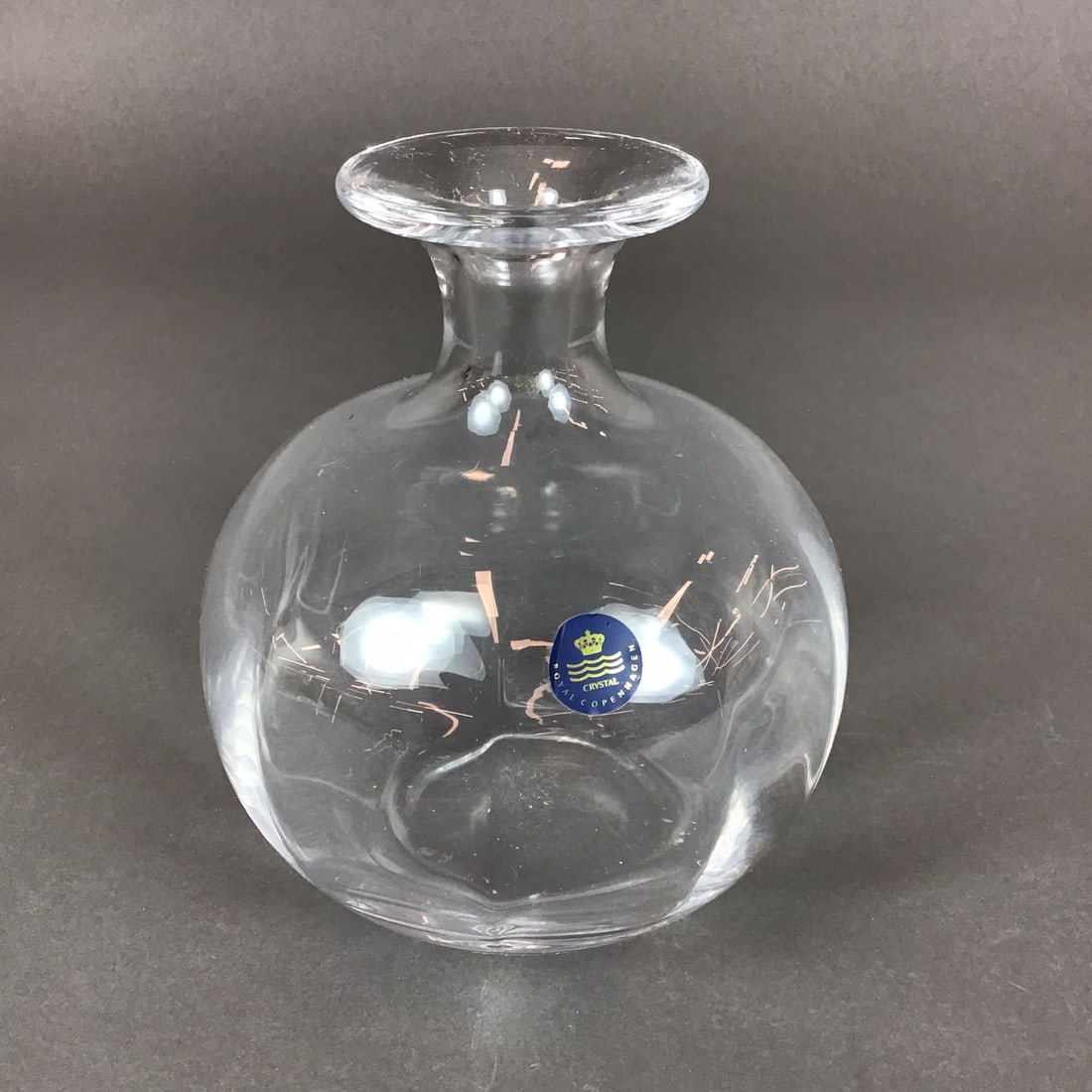 Royal Copenhagen Crystal Decanter: This medium-sized crystal decanter is produced by Royal Copenhagen. The piece is crafted from clear crystal and features a bulbous, spherical body that transitions into a narrow, tapered neck with a w