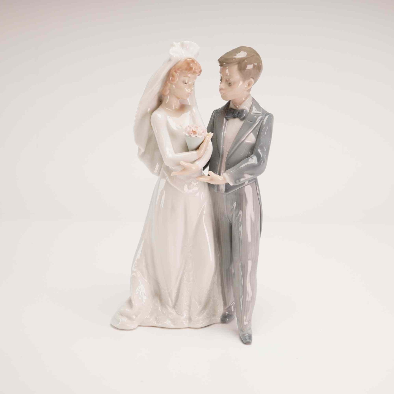 Lladro Glazed Porcelain Figurine From This Day Forward 01005885 with Box: The piece depicts a bride and groom in formal wedding attire. The bride is dressed in a flowing white gown with a long veil and holds a small bouquet of pink flowers. The groom stands alongside her in