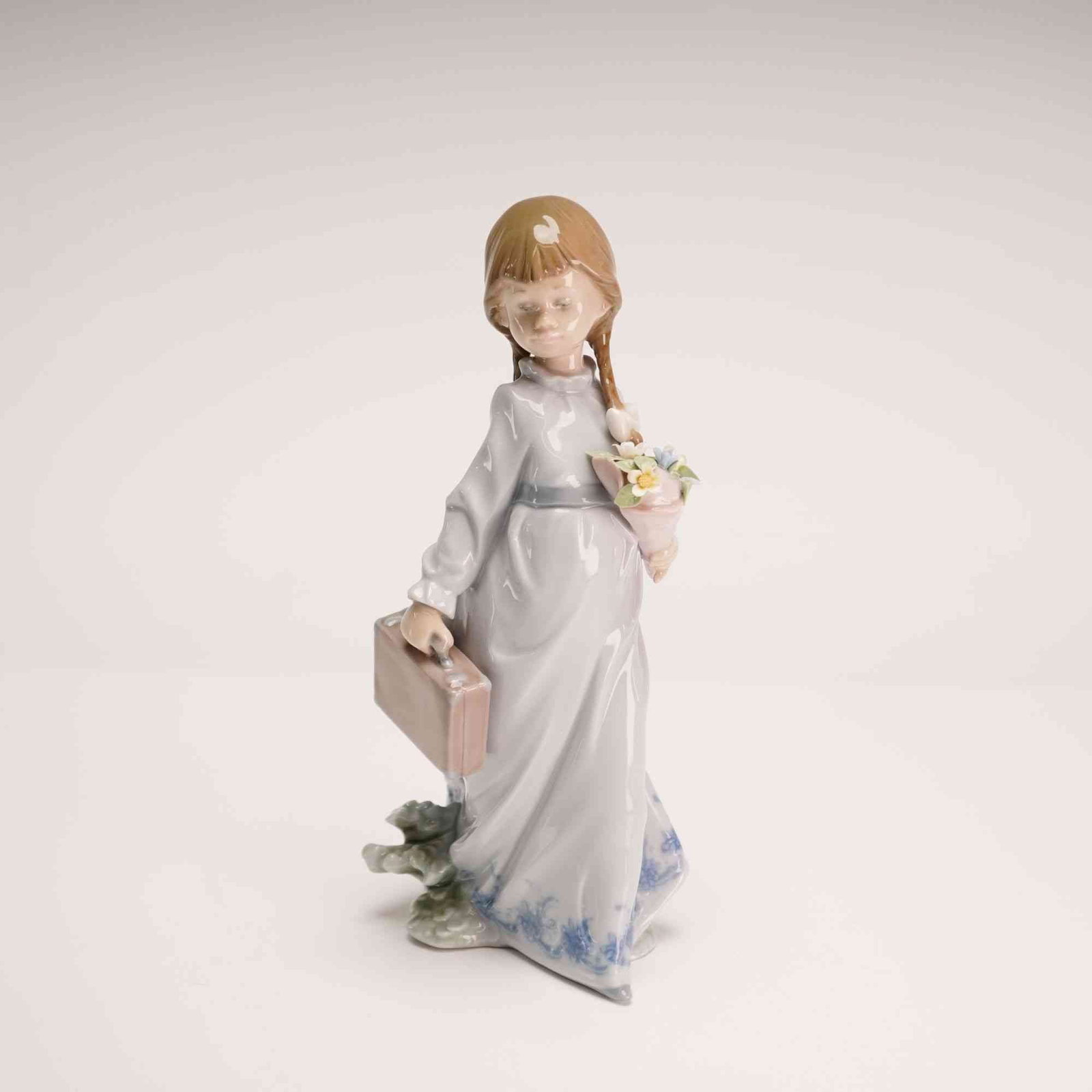 Lladro Porcelain Figurine School Days 01007604: The sculpture, designed by Antonio Ramos, depicts a young girl with blonde braids wearing a pale blue gown with a darker blue sash and floral-patterned trim along the hem. She is portrayed carrying a 