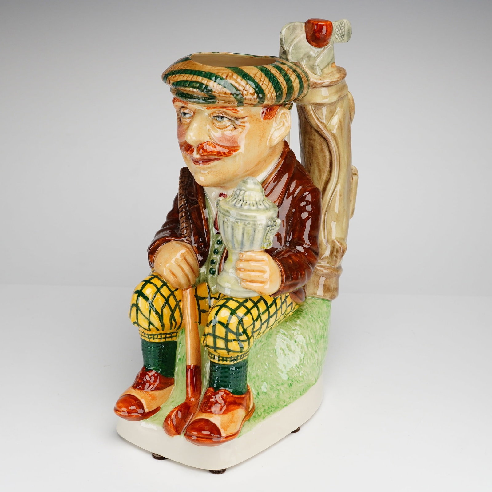 Kevin Francis Ceramics Limited Edition Toby Jug, The Golfer with COA: The molded ceramic figure depicts a seated golfer in traditional attire, including a green and tan striped flat cap, a brown jacket over a green vest and red tie, and yellow plaid knickerbockers. The
