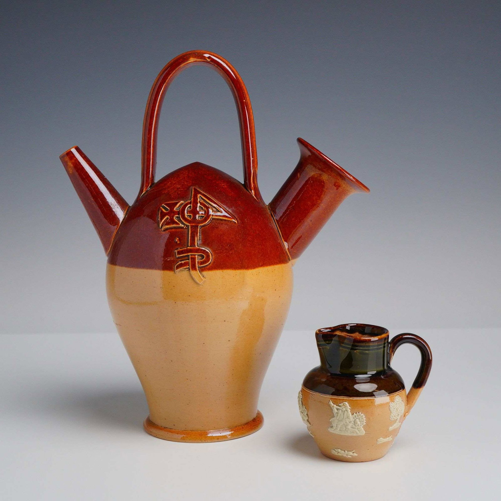 2pc Royal Doulton Lambeth Stoneware Items, The Old Sarum Kettle & Creamer w/ Reliefs: This lot consists of two pieces of Doulton Lambeth salt-glazed stoneware featuring traditional two-toned finishes and applied relief decoration. The primary item is known as The Old Sarum Kettle, a