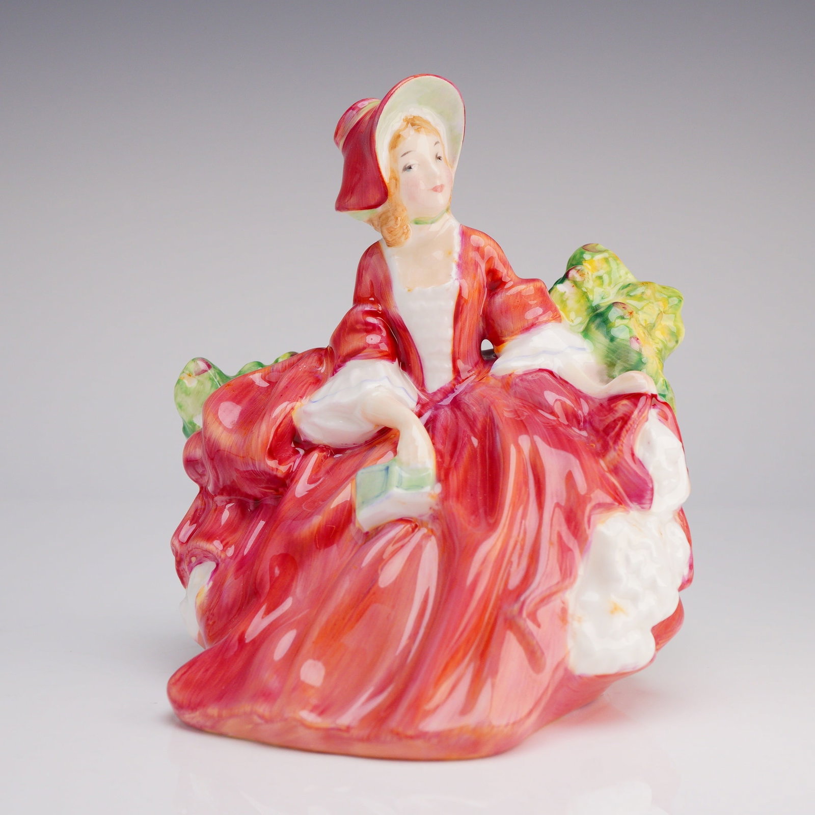 Royal Doulton Figurine Lydia HN 1908: The figure depicts a seated young woman wearing a wide-brimmed bonnet and a voluminous period dress primarily rendered in vibrant red and white glazes, resting against green and yellow foliage. Web re