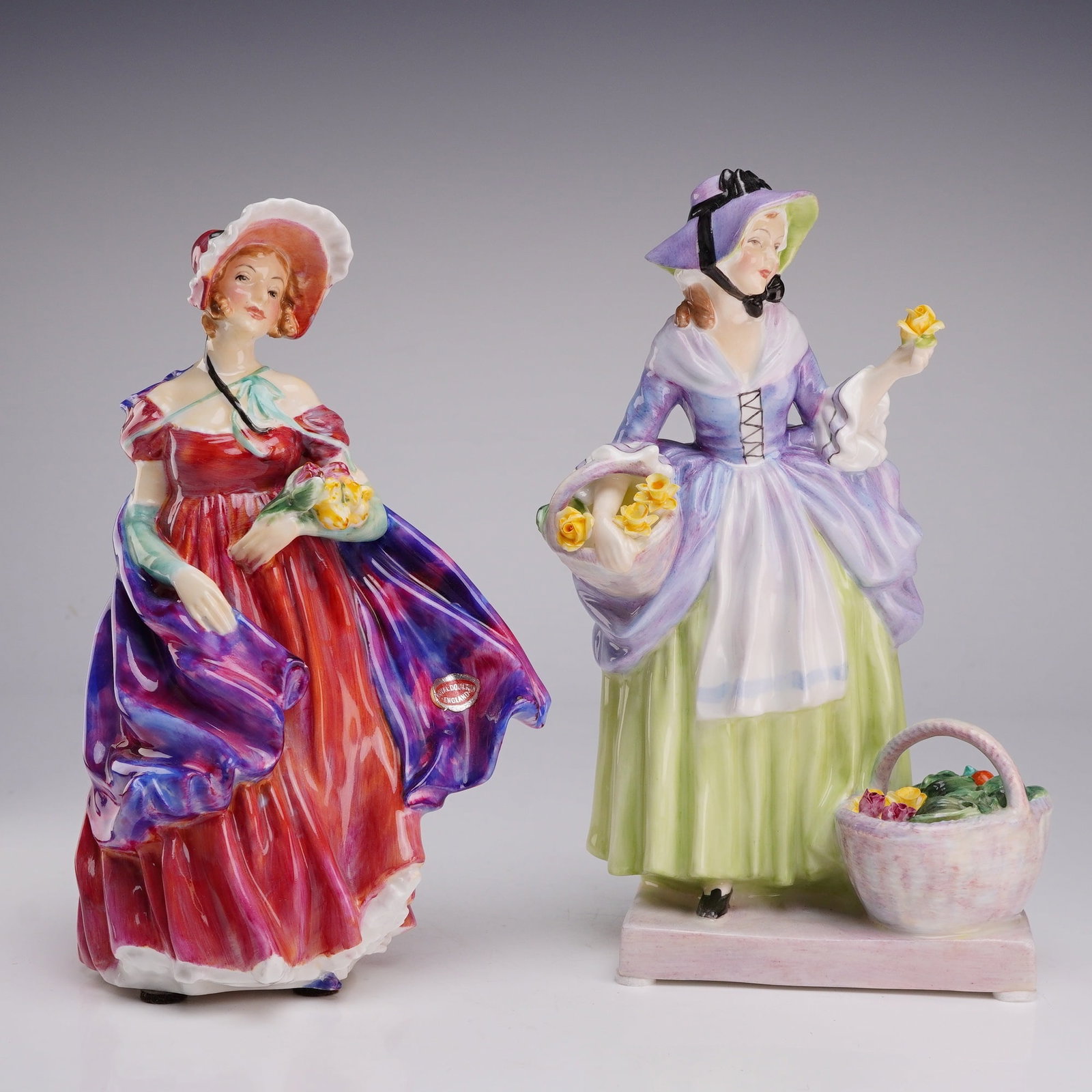 Two Royal Doulton Bone China Figurines, Lady April HN1958 and Springflowers HN1807: The first figurine is titled Lady April, model number HN1958. It depicts a standing female figure in a red dress with a voluminous shawl featuring mottled purple and blue glazes. She wears a large whi