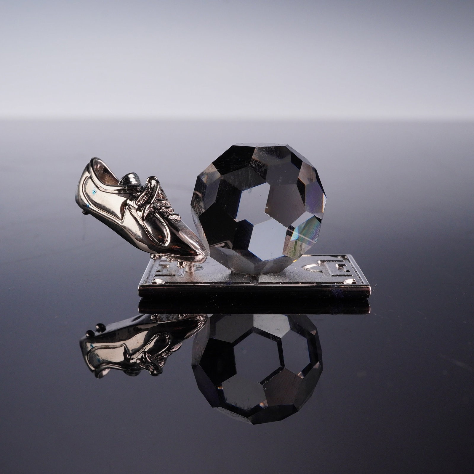 Swarovski Crystal Memories Figurine, Rhodium Soccer Trophy: This miniature figurine features a faceted clear crystal soccer ball and a rhodium-plated silver-tone metal soccer boot positioned in a kicking motion. The components are mounted on a rectangular silv