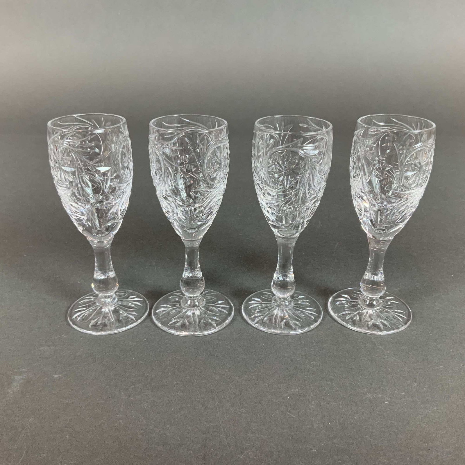 A Set of Four Tudor England Crystal Liqueur Glasses: A set of four Tudor England crystal liqueur glasses. Each glass features an intricate wheel-cut pattern on the bowl consisting of scrolling foliate and floral motifs interspersed with star rosettes.