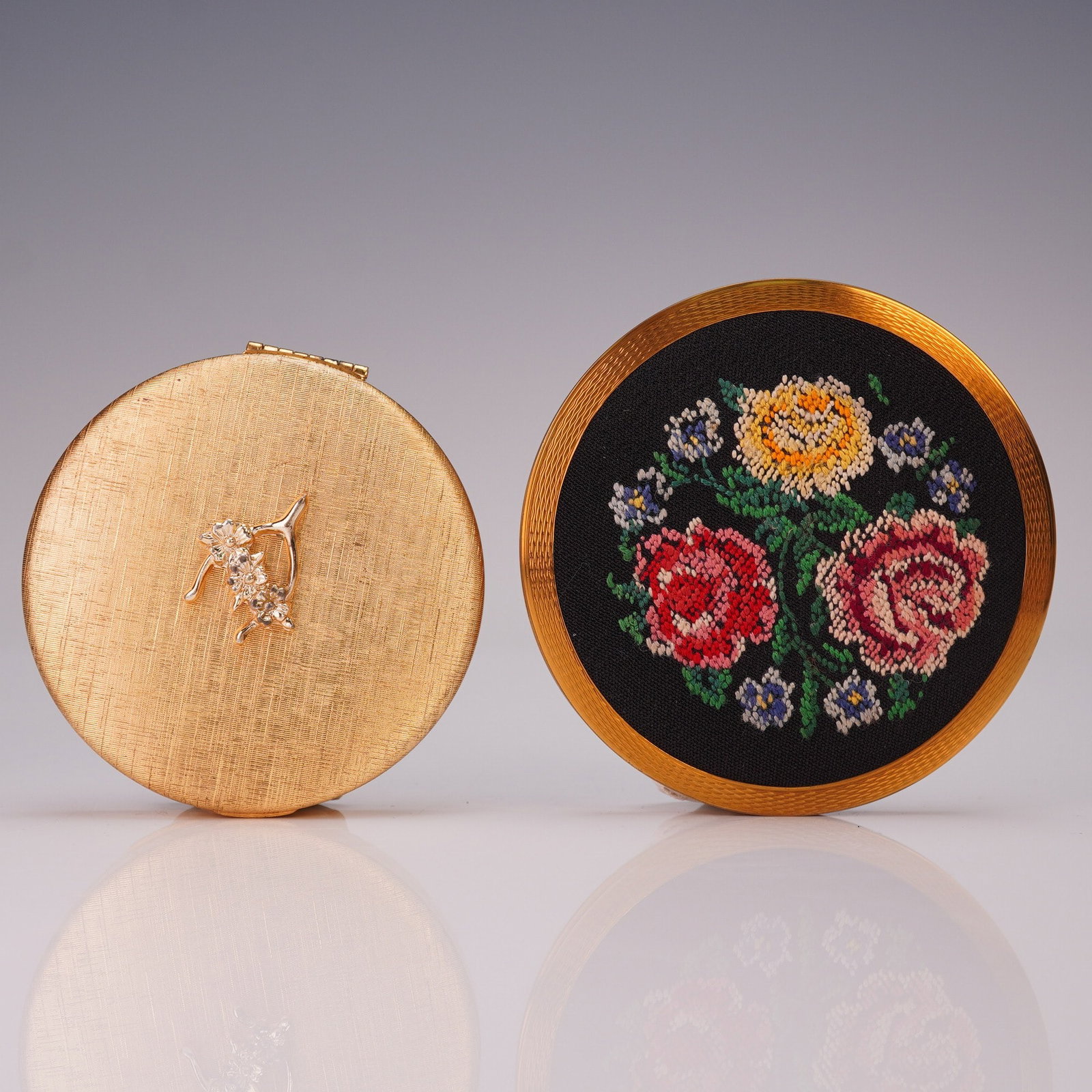 2pc Vintage Stratton and Dubarry Compact Cases: Pair of distinctive round compacts including a Stratton England compact with gold-tone casing, black enamel back, and floral petit point embroidery on the lid, and a Dubarry CloudSilk compact in gold-