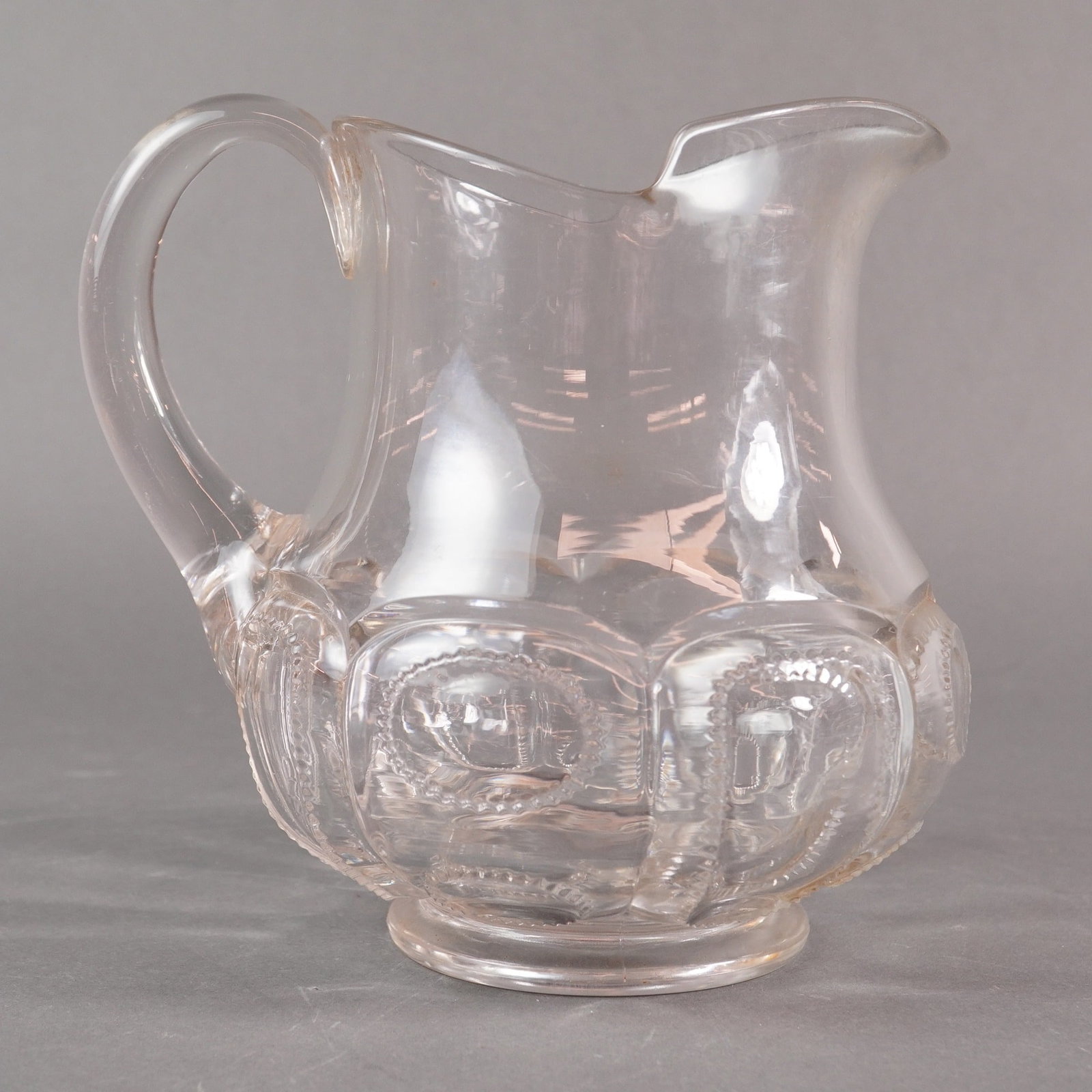 An Early American Pressed Glass (EAPG) Clear Medallion Pattern Pitcher: A single Early American Pressed Glass (EAPG) clear glass pitcher. The vessel features a bulbous lower body that transitions to a narrower neck, complete with a pronounced pouring spout and an