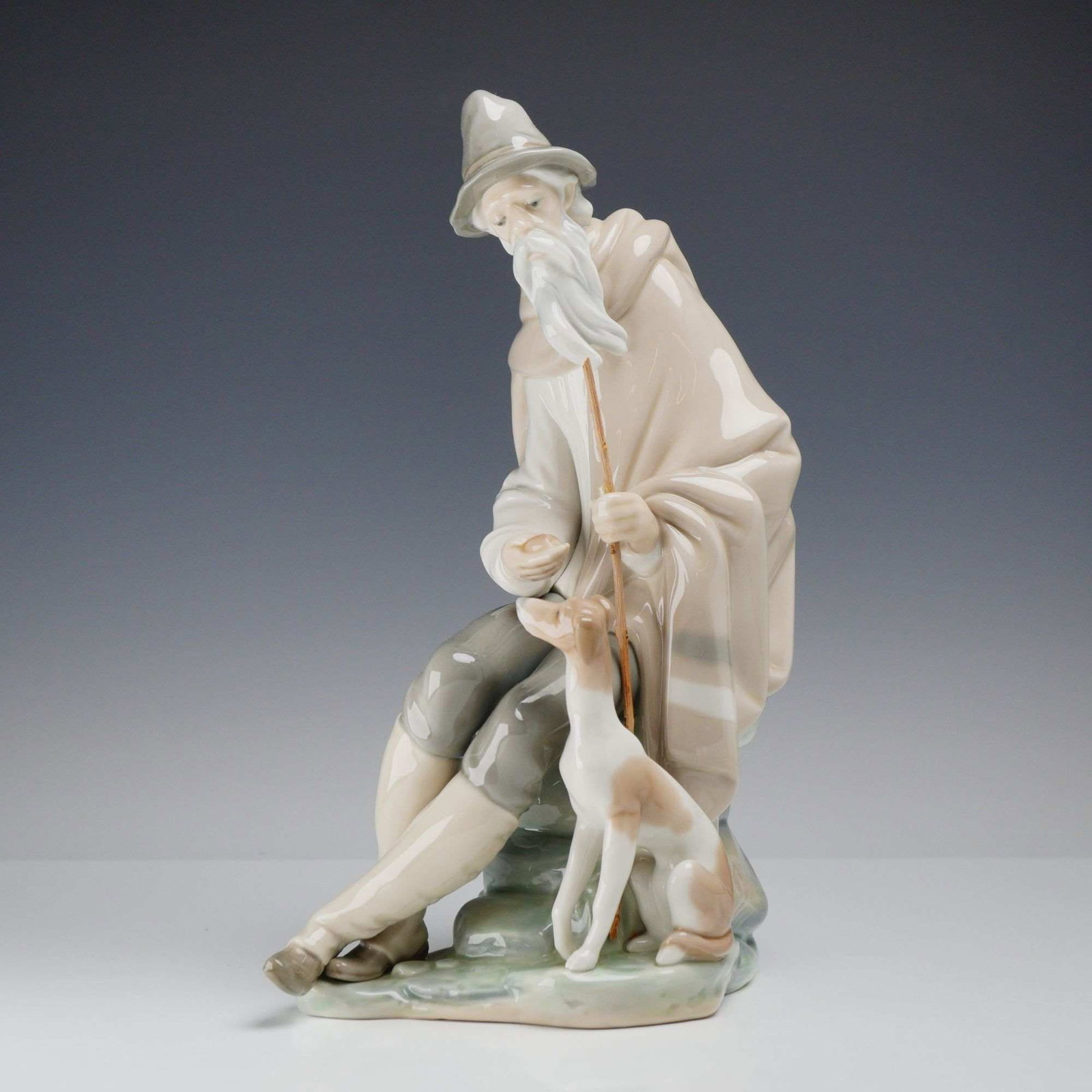 Lladro Beggar Seated with Dog Porcelain Figurine Model 01001094: A glazed porcelain figurine manufactured by Lladro in Spain, titled Beggar (Mendigo), item number 01001094. Designed by artist Juan Huerta, the piece depicts an elderly seated man with a flowing
