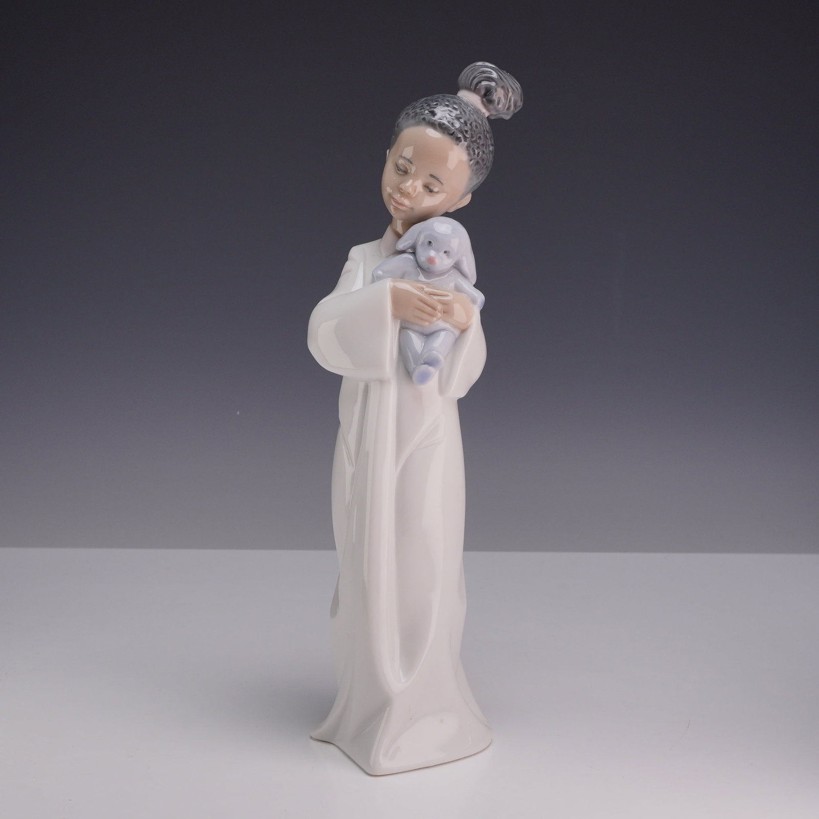 Lladro Black Porcelain Figurine, My Cuddly Puppy 1006463 with Original Box: The figurine depicts a young Black girl wearing a long white bathrobe with draped sleeves, cradling a pale blue stuffed bunny. The figure is modeled with a neutral facial expression and hair styled