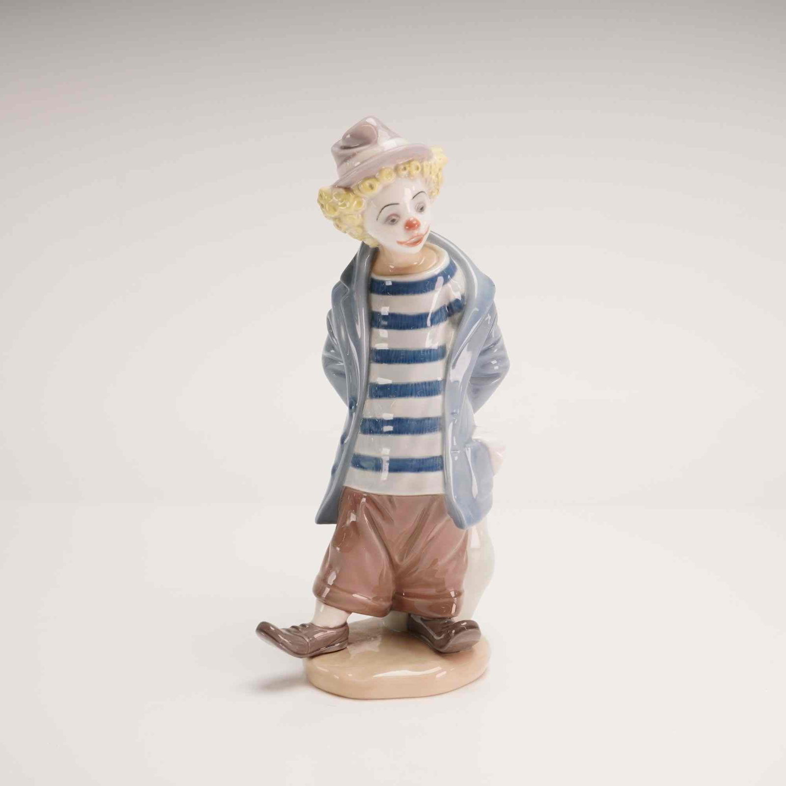 Lladro Porcelain 1986 Collectors Society Figurine Little Traveler 01007602: This glazed porcelain figurine, was designed by Regino Torrijos for Lladro. The figure depicts a young clown in a hobo style, standing with his hands behind his back holding a white sack or bundle.