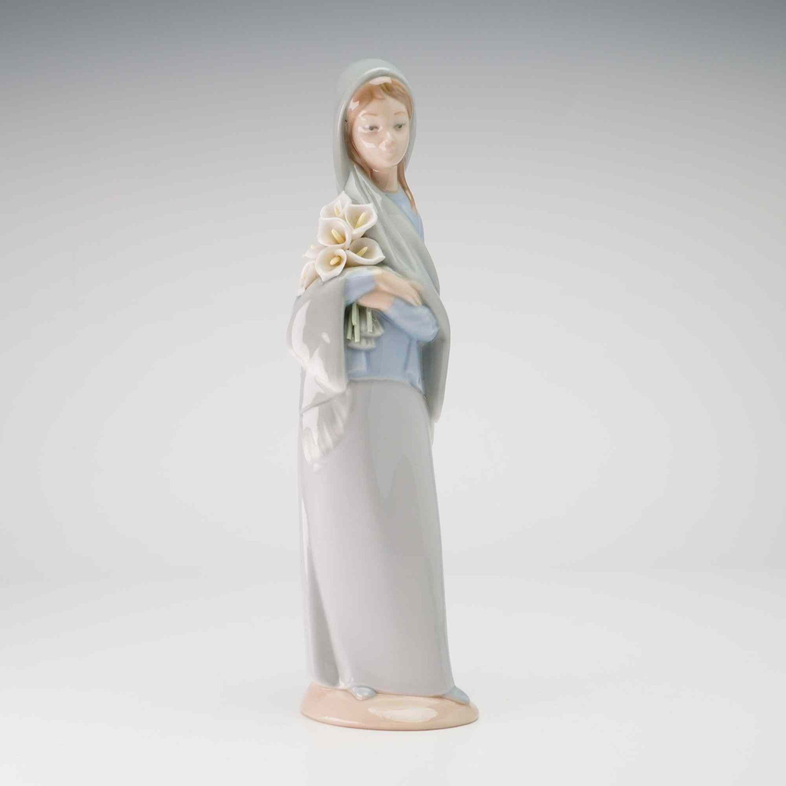 Lladro Porcelain Figurine Girl with Calla Lilies 01004650: The figurine depicts a standing young woman dressed in a pale blue bodice and a long gray skirt, with a matching gray veil or headscarf draped over her head and shoulders. She holds a cluster of white