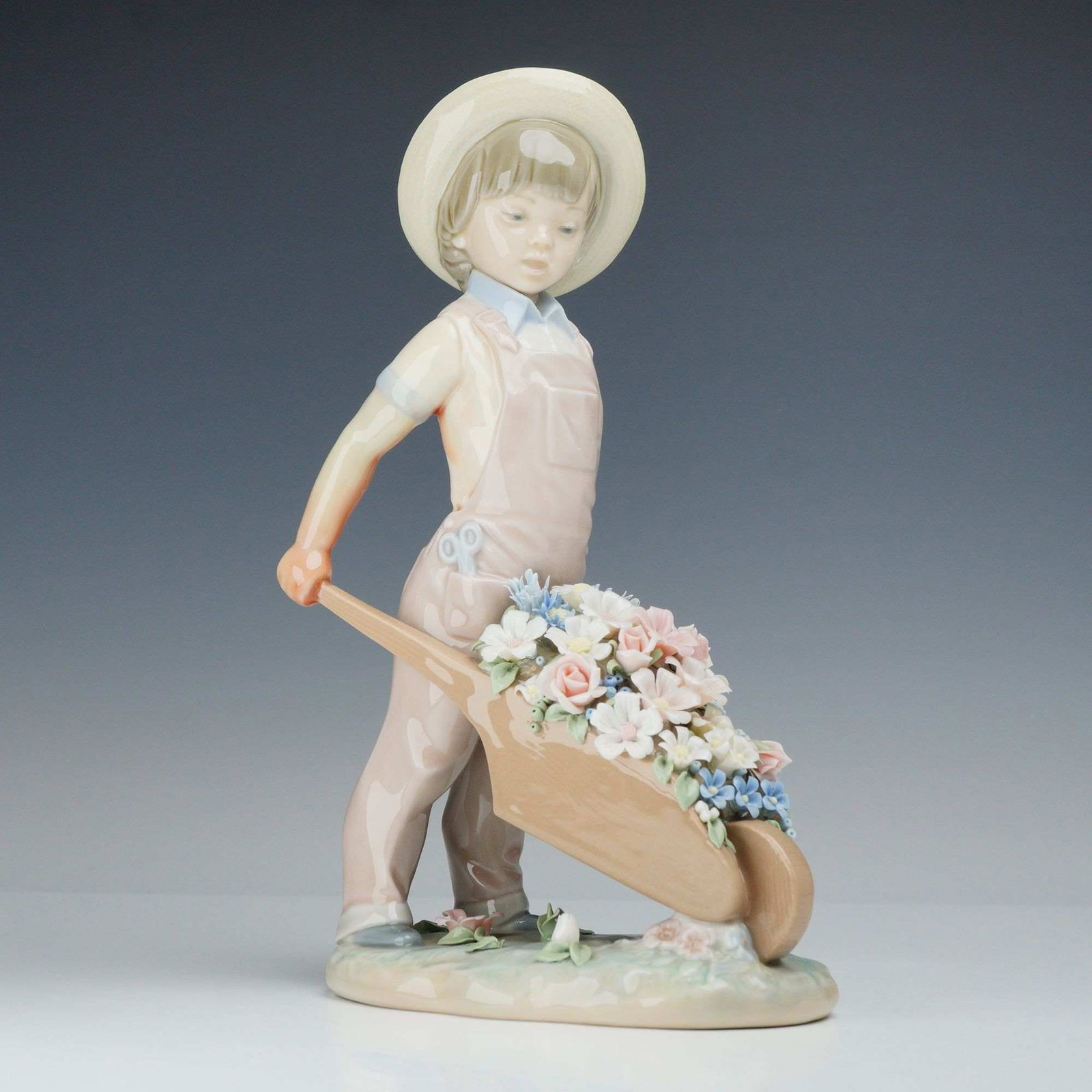 Lladro Porcelain Figurine Little Gardener Number 01001283: A glazed porcelain figurine titled Little Gardener (Spanish title: Carretilla con Flores), model number 01001283, produced by Lladro in Spain. Sculpted by Francisco Catala, this piece depicts a young 