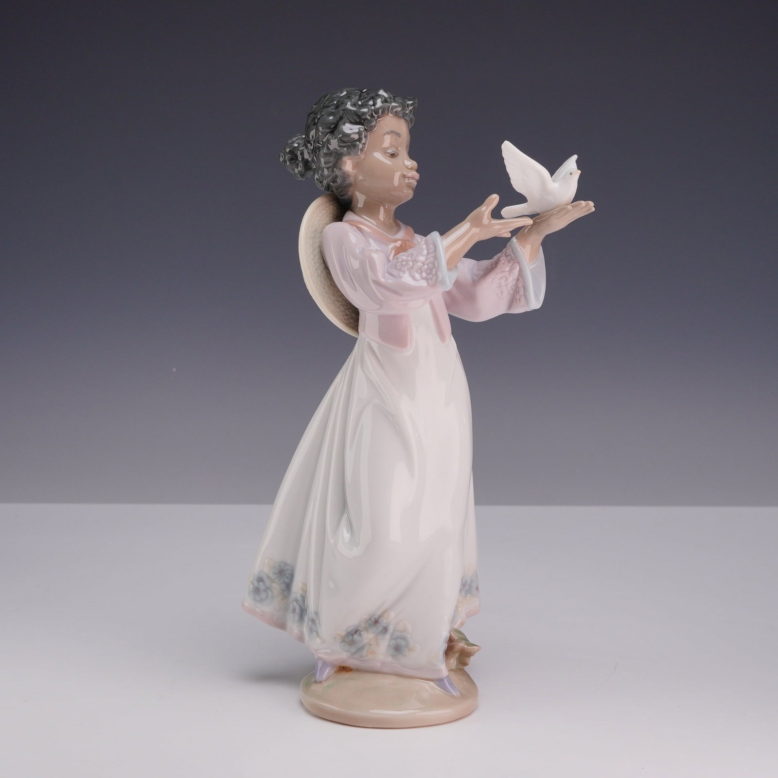 Lladro Porcelain Figurine, African Love 1006154 with Box: The figurine depicts a young girl with a dark complexion and curly black hair gathered in a bun. She is dressed in a pale pink jacket over a long white gown decorated with a blue and lavender floral p