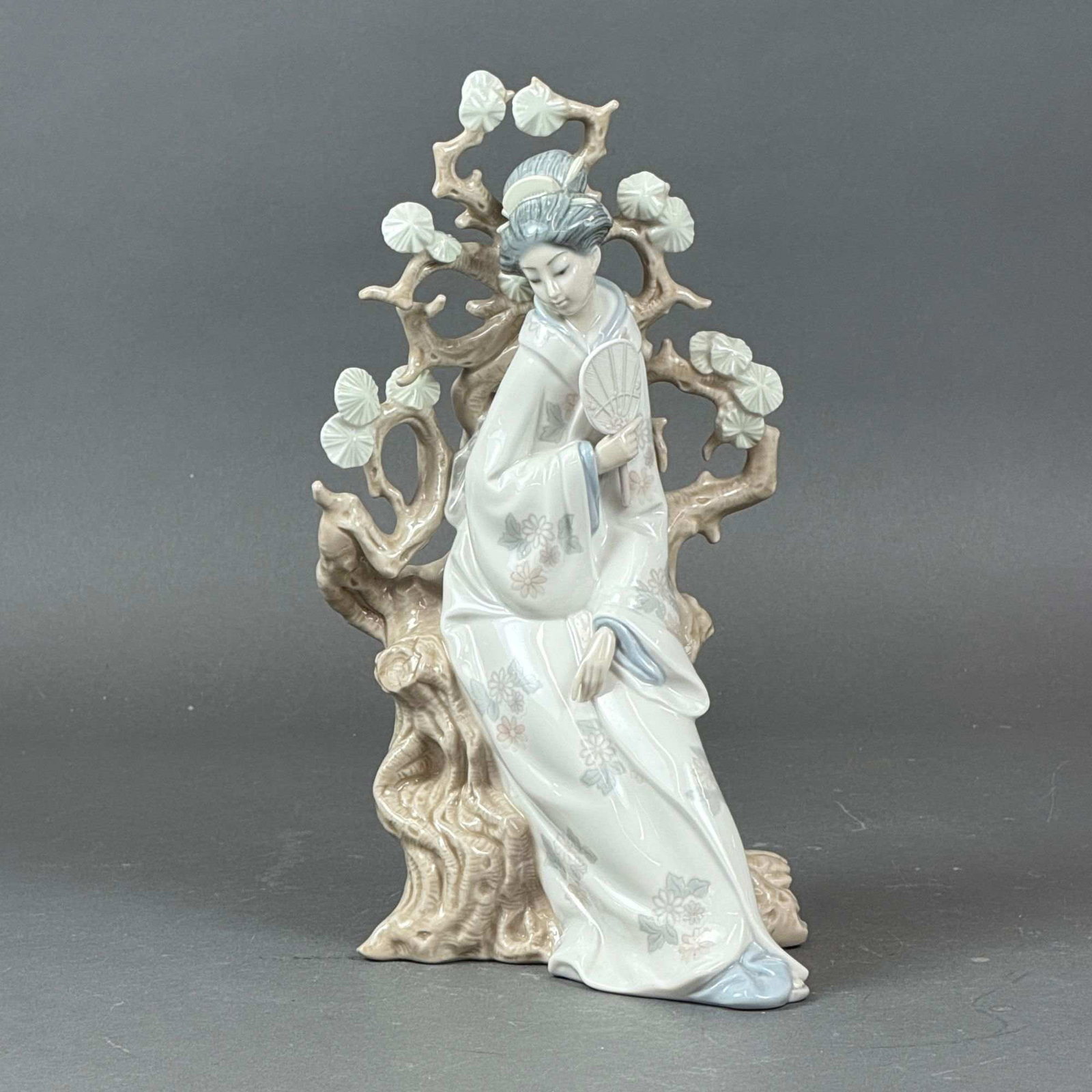 Lladro Porcelain Figurine, Geisha 01004807: Porcelain sculpture depicts a serene geisha figure seated gracefully before an intricately sculpted tree, her flowing kimono adorned with delicate floral patterns. The composition beautifully highligh