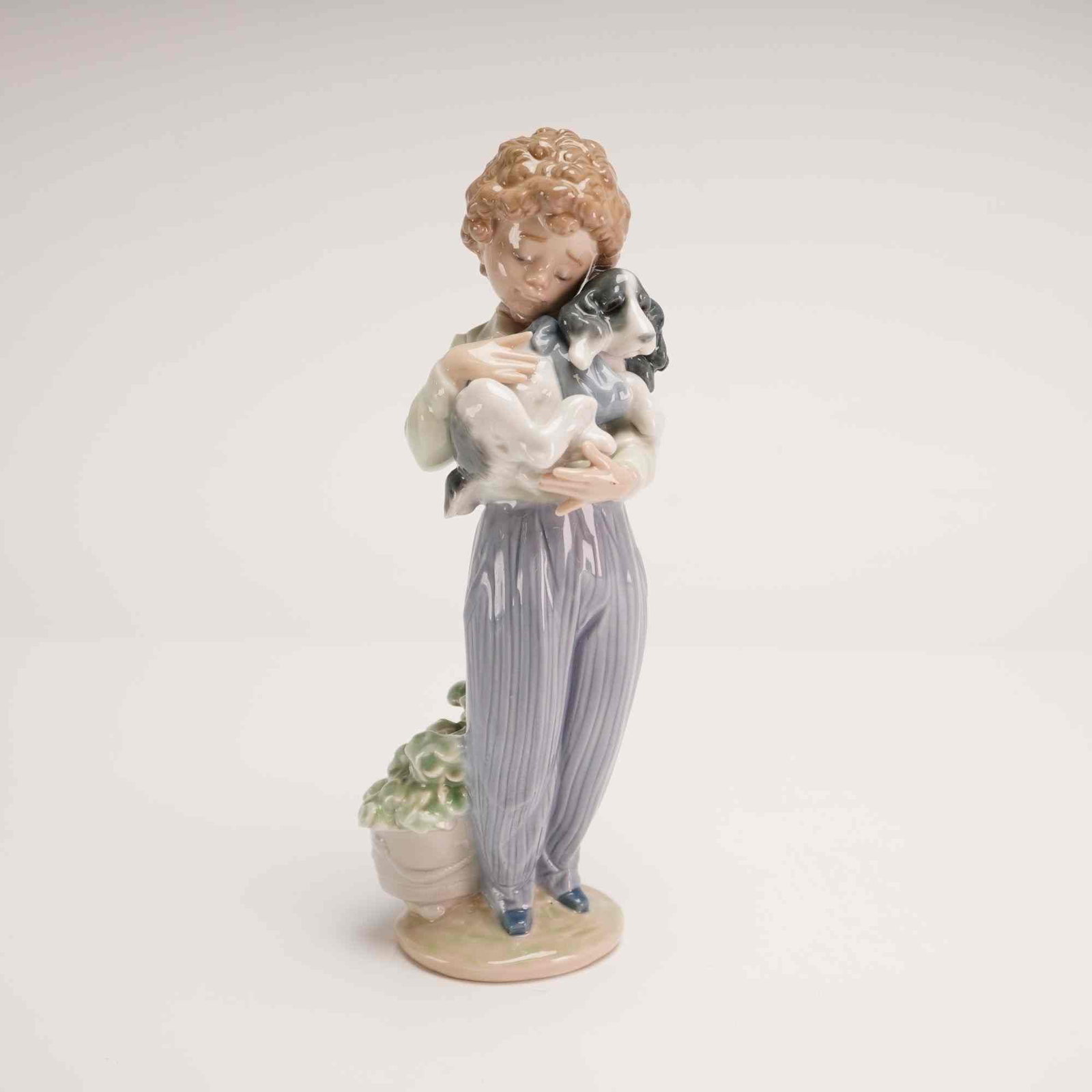 Lladro Porcelain Figurine, My Buddy 01007609: Glazed porcelain figurine issued for the 1989 Collectors Society. The sculpture depicts a young boy with curly brown hair holding a spaniel puppy. The figure is dressed in lavender-striped trousers an