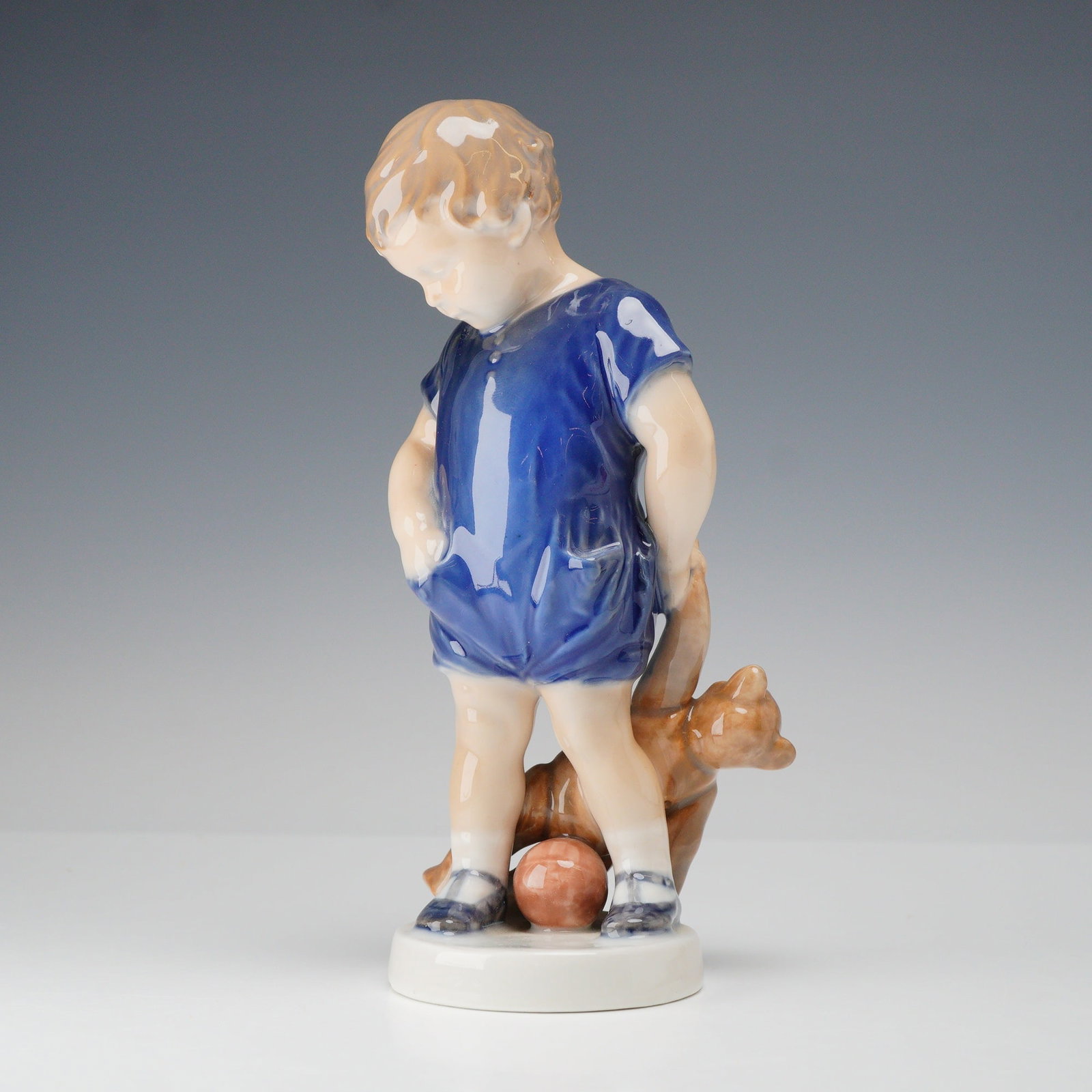 Royal Copenhagen Porcelain Figurine, Boy with Teddy Bear 3468: A Royal Copenhagen glazed porcelain figurine depicting a young boy wearing a blue romper. The child is depicted standing with his right hand in a pocket and his left hand behind his back, holding a br