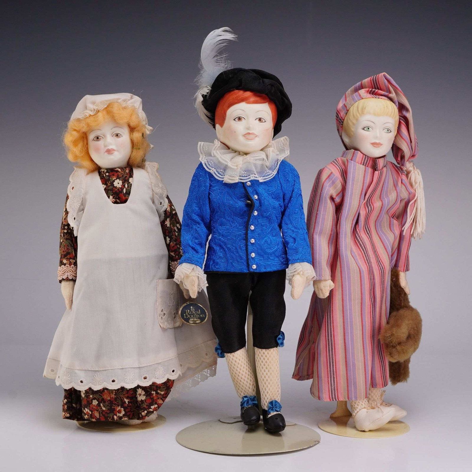 3pc Royal Doulton Victorian Birthday Doll Collection in Porcelain: A trio of dolls from the Royal Doulton Victorian Birthday Doll Collection. Each doll is designed with a bisque porcelain head and hands joined to a stuffed cloth body. The collection includes a girl w