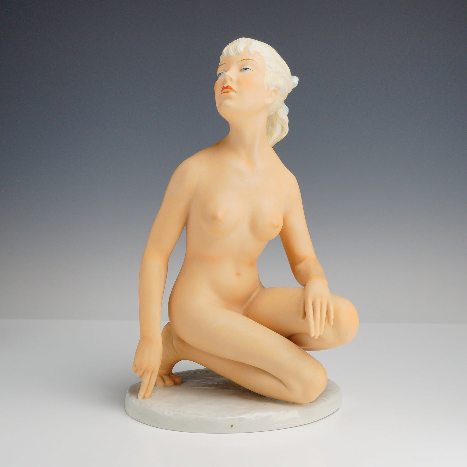 H. Schreiber Porcelain Figurine of a Naked Woman by K. Steiner: This hand-painted figure is depicted in a kneeling pose with a slight twist to the torso, gazing upward and to her left. The body features a matte bisque finish to simulate skin texture, while the blo