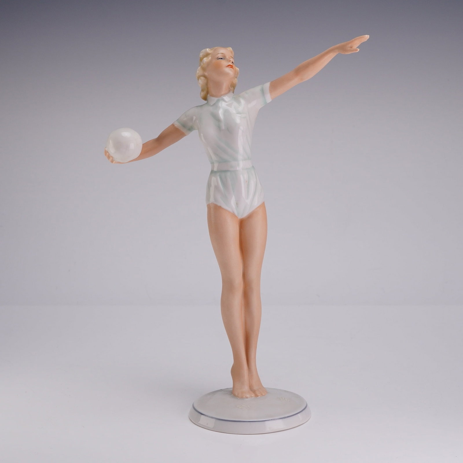 Schaubach Kunst Art Deco Porcelain Volleyball Player Figurine: This porcelain figurine, produced by Schaubach Kunst in Germany, depicts a female athlete in an Art Deco style. The figure is captured in a graceful pose, holding a white ball in her right hand with h