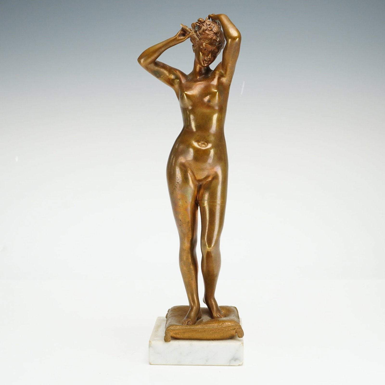 Art Nouveau Bronze Sculpture, Female Nude Combing her Hair: This bronze sculpture depicts a standing female nude shown in the act of arranging her hair with both arms raised. The figure is positioned atop a rectangular bronze tasseled cushion, which is further
