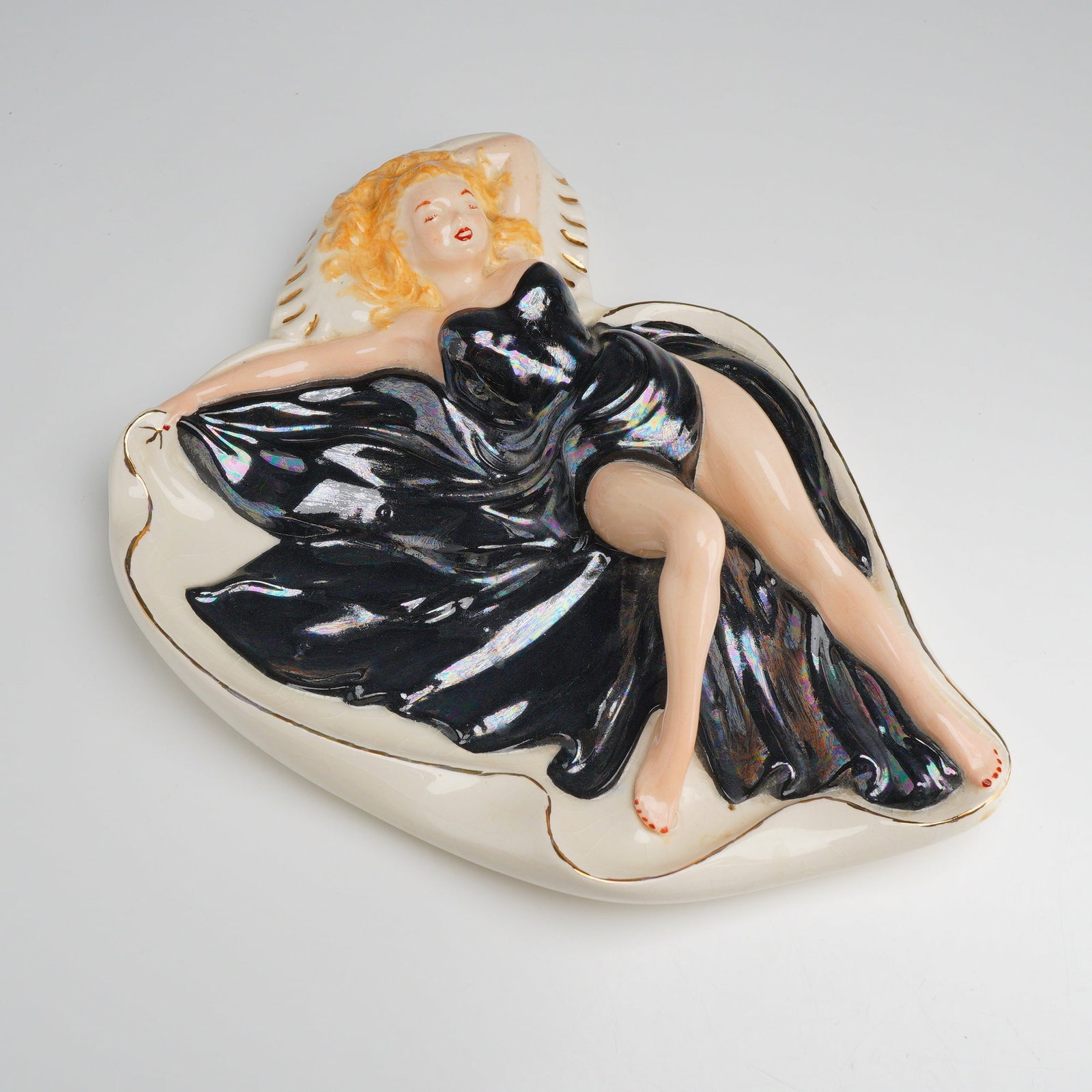 Mid-Century Ceramic Pin-Up Style Girl in Black Ashtray: This mid-century modern pottery figurine depicts a reclining female figure in a pin-up pose. The figure is dressed in a strapless black gown featuring an iridescent luster finish. She is positioned