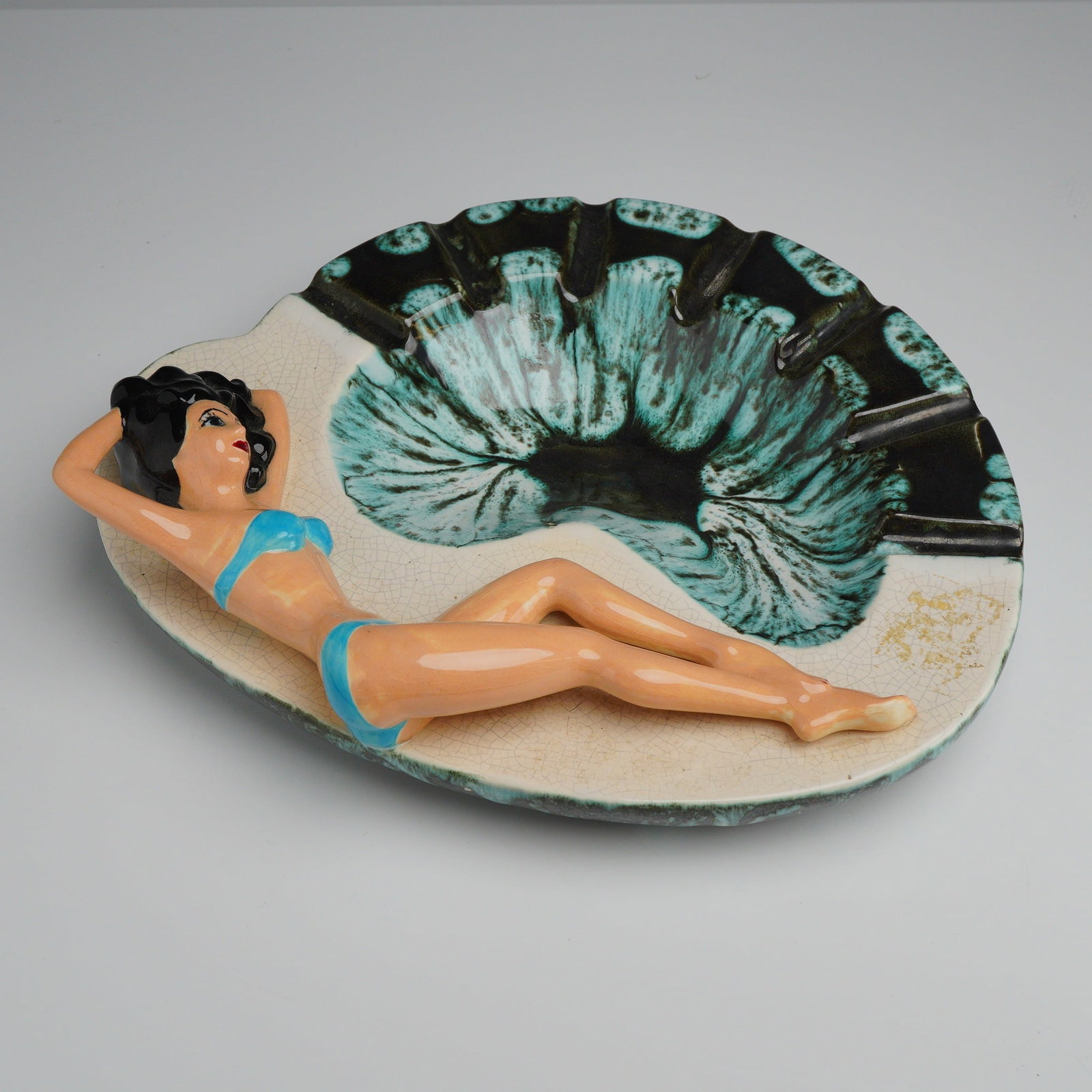Mid-Century Ceramic Pin-up Style Girl in Blue Bikini Ashtray: This vintage ceramic woman ashtray is a mid-century novelty piece produced in the style of California Pottery from the 1950s. The item is constructed from glazed earthenware in a freeform, shell-like 