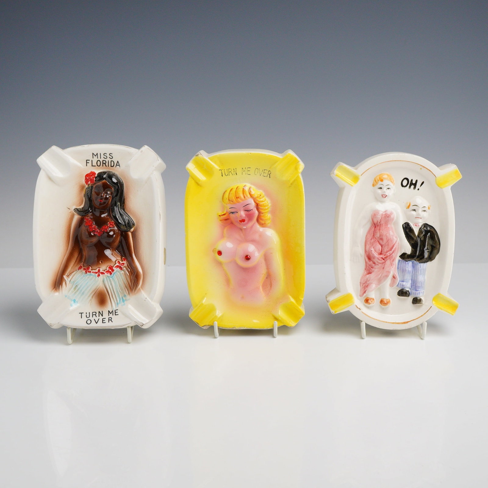 Three Risque Novelty Ceramic Ashtrays: A collection of four mid-century novelty porcelain ashtrays featuring risque relief-molded figures and humorous text. The group includes a white rectangular ashtray depicting a woman in a lei and