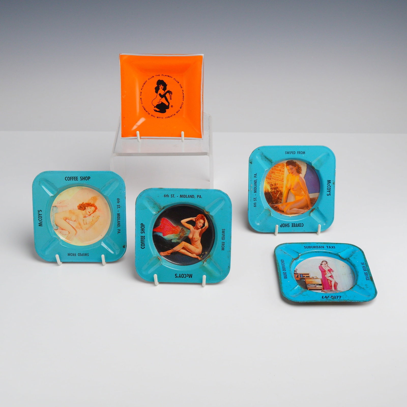 Five Vintage Risque Advertising Pin-Up Ashtrays: A group of five vintage advertising ashtrays featuring pin-up imagery. The collection includes one square orange plastic ashtray and four lithographed tin ashtrays. The plastic example is decorated